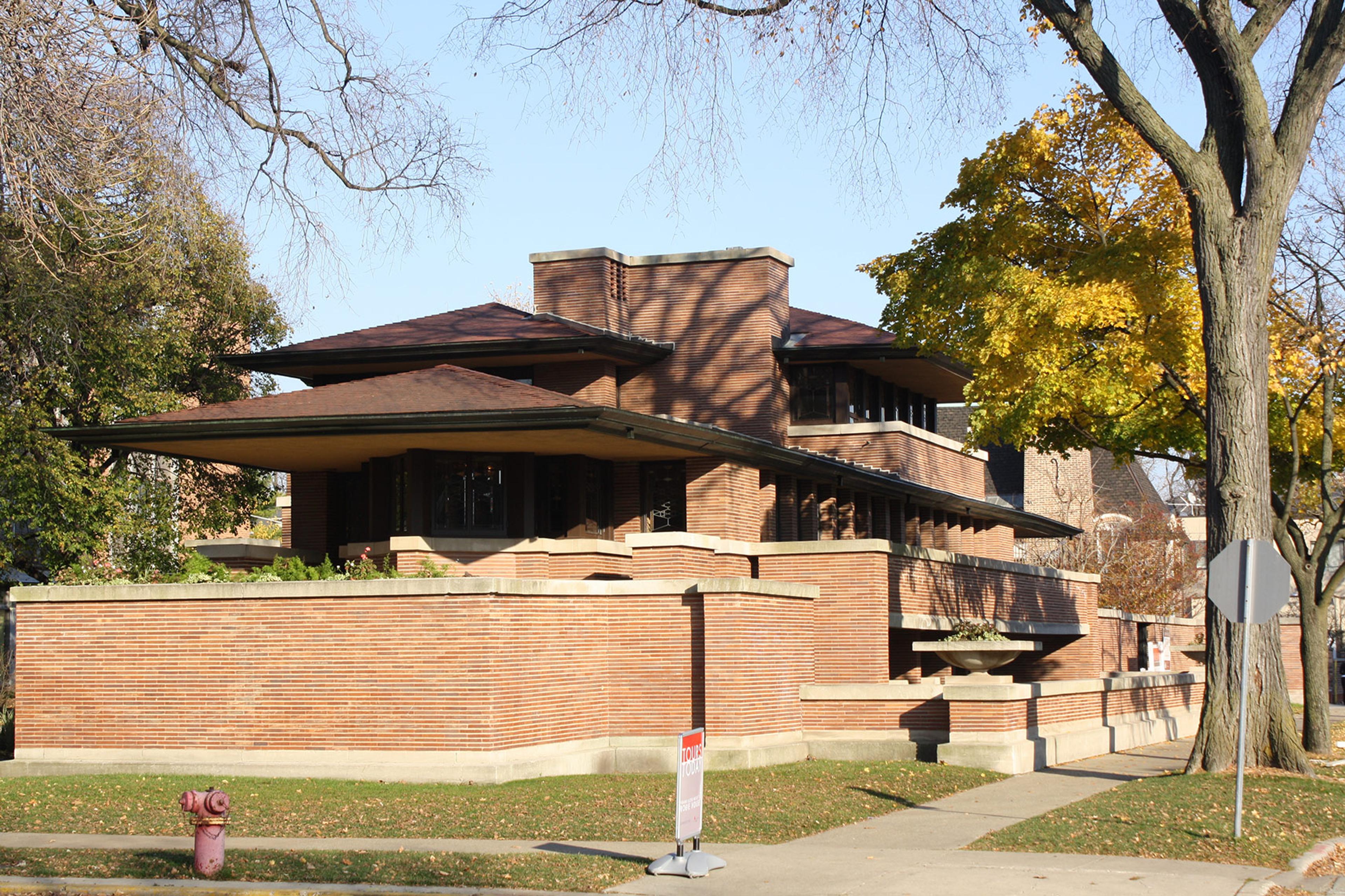 A brick house with a pitched roofs and large eaves surrounded by trees, taken on a sunny day.