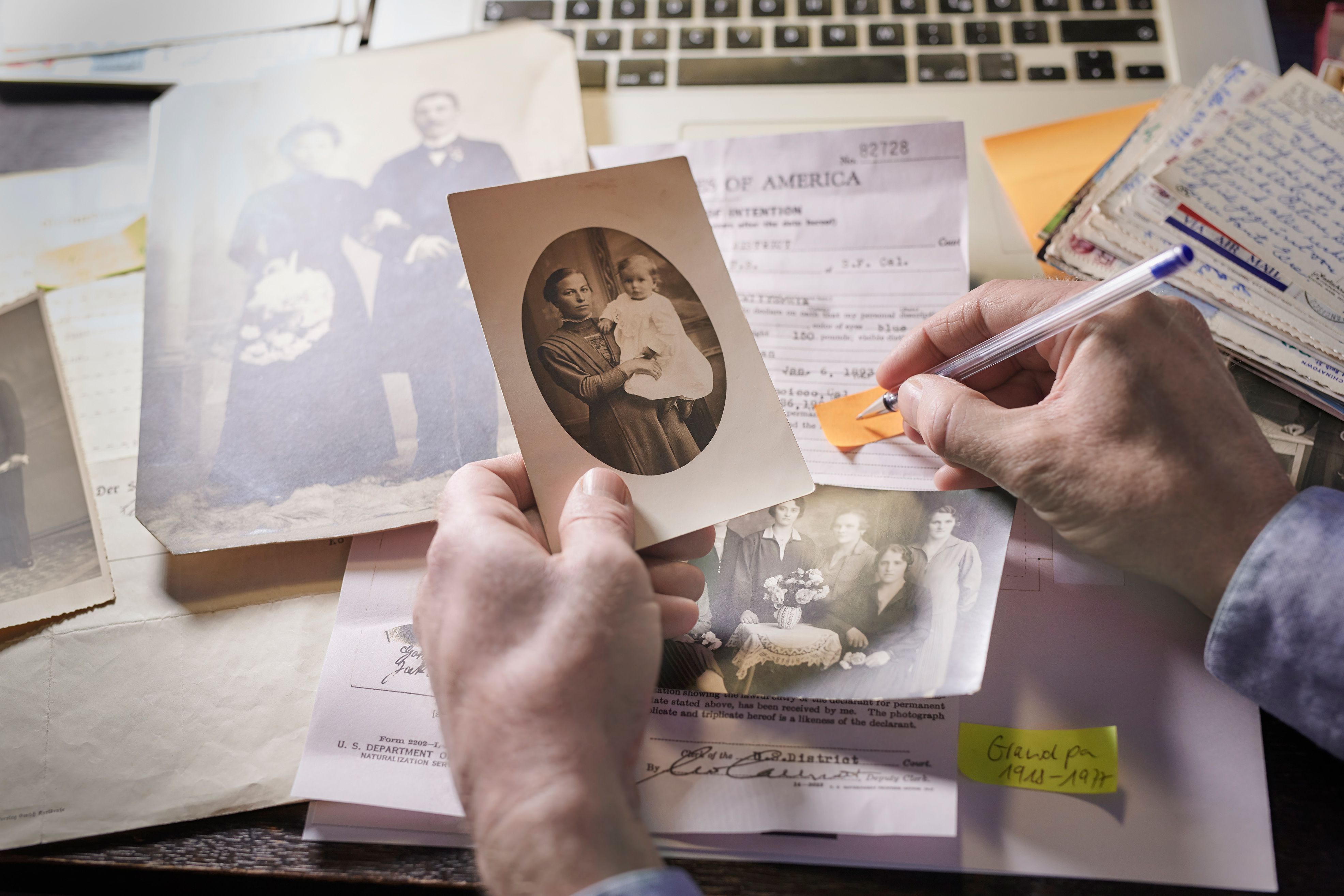 A person’s left hand holds an old family photo near documents and letters on a table; their right hand holds a pen that is positioned to write on a yellow strip of paper.