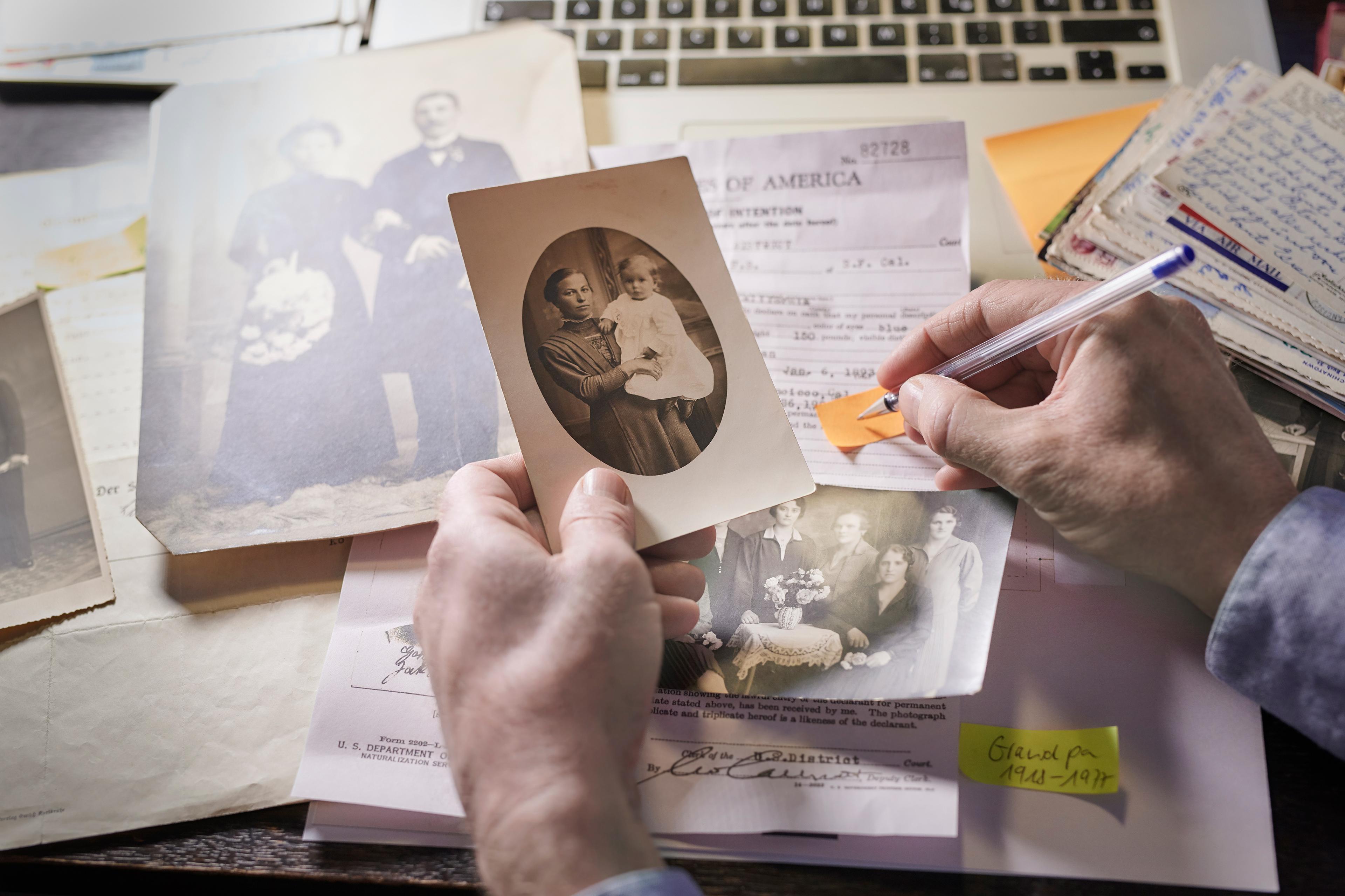 A person’s left hand holds an old family photo near documents and letters on a table; their right hand holds a pen that is positioned to write on a yellow strip of paper.