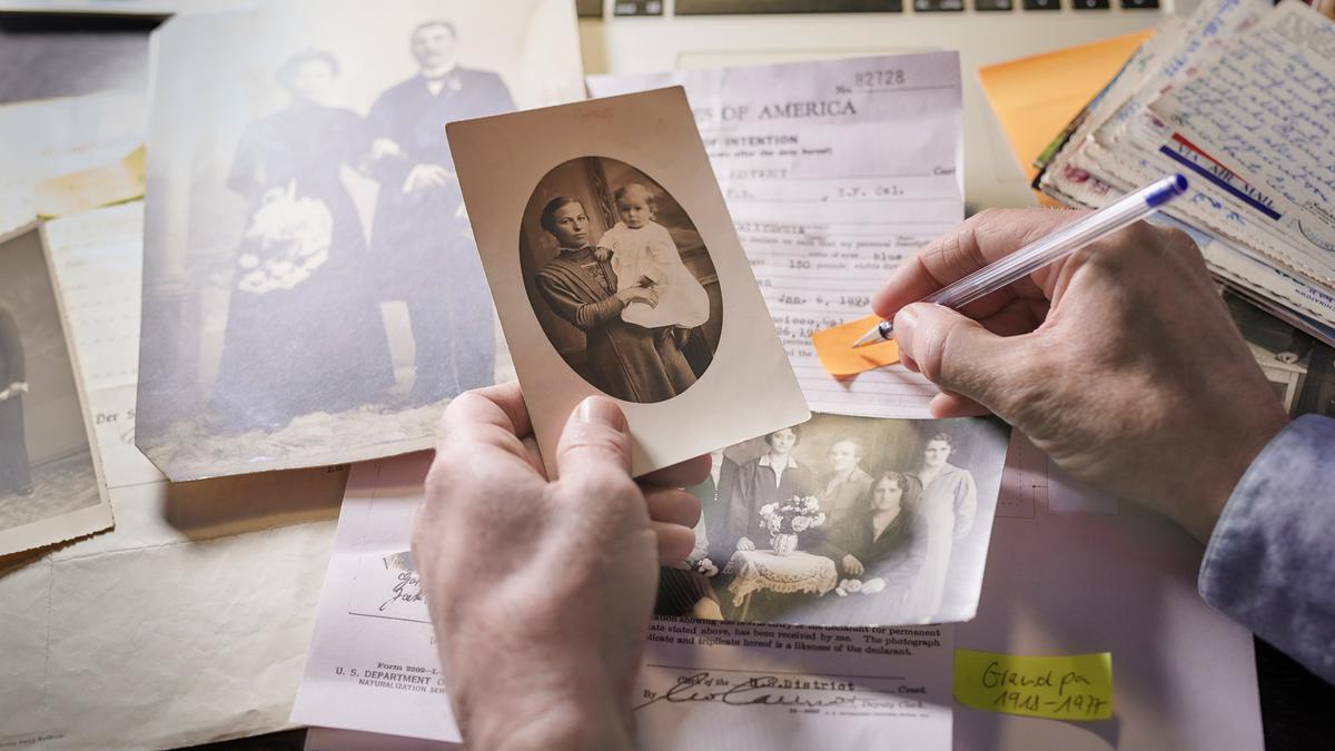 A person’s left hand holds an old family photo near documents and letters on a table; their right hand holds a pen that is positioned to write on a yellow strip of paper.