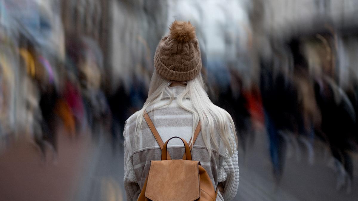 A person with long blonde hair in a beanie and coat, facing away from the camera, wearing a brown backpack in a blurred busy street.