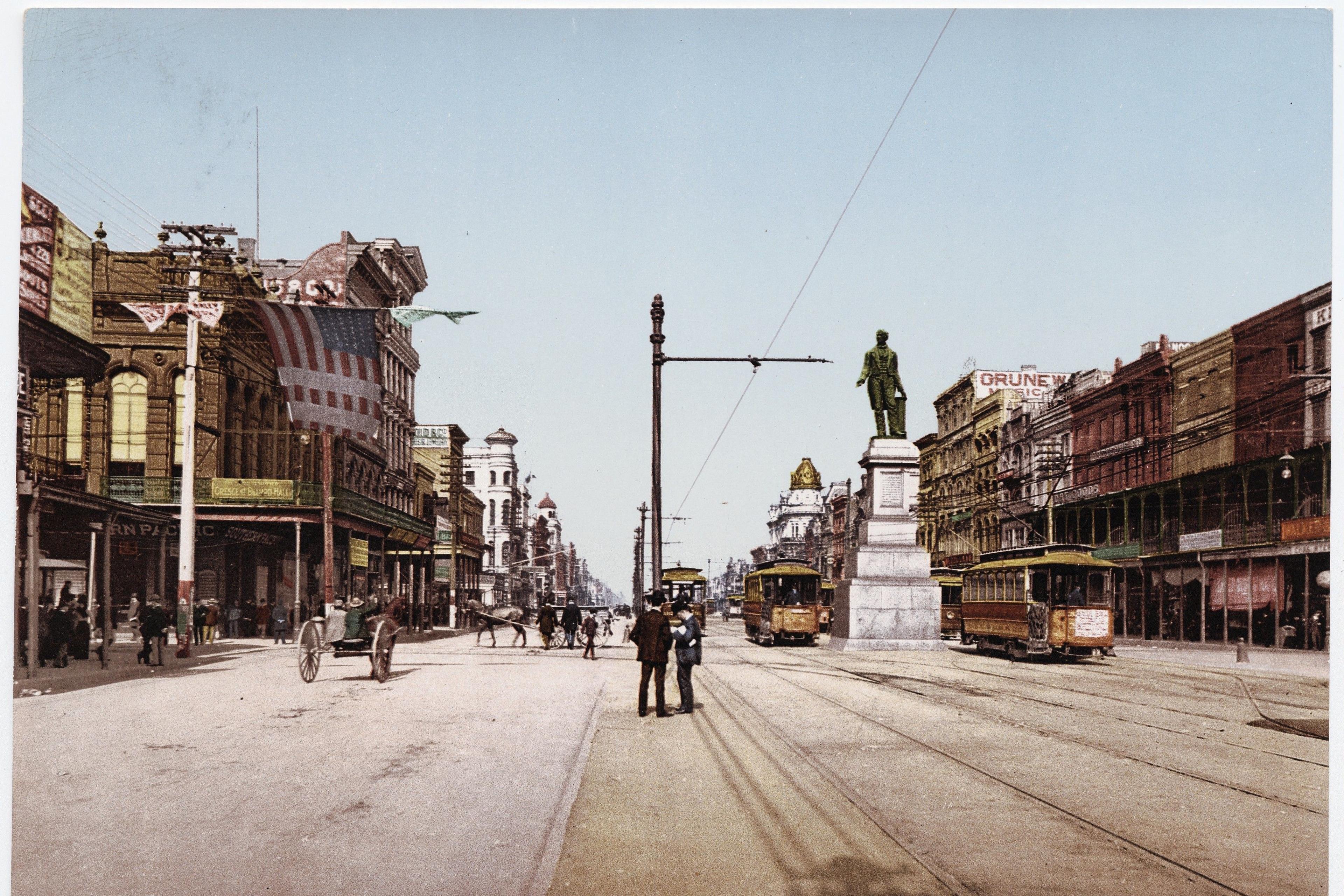 Hand-coloured photo of Canal Street New Orleans featuring trams, a statue and historic buildings in early 20th century.