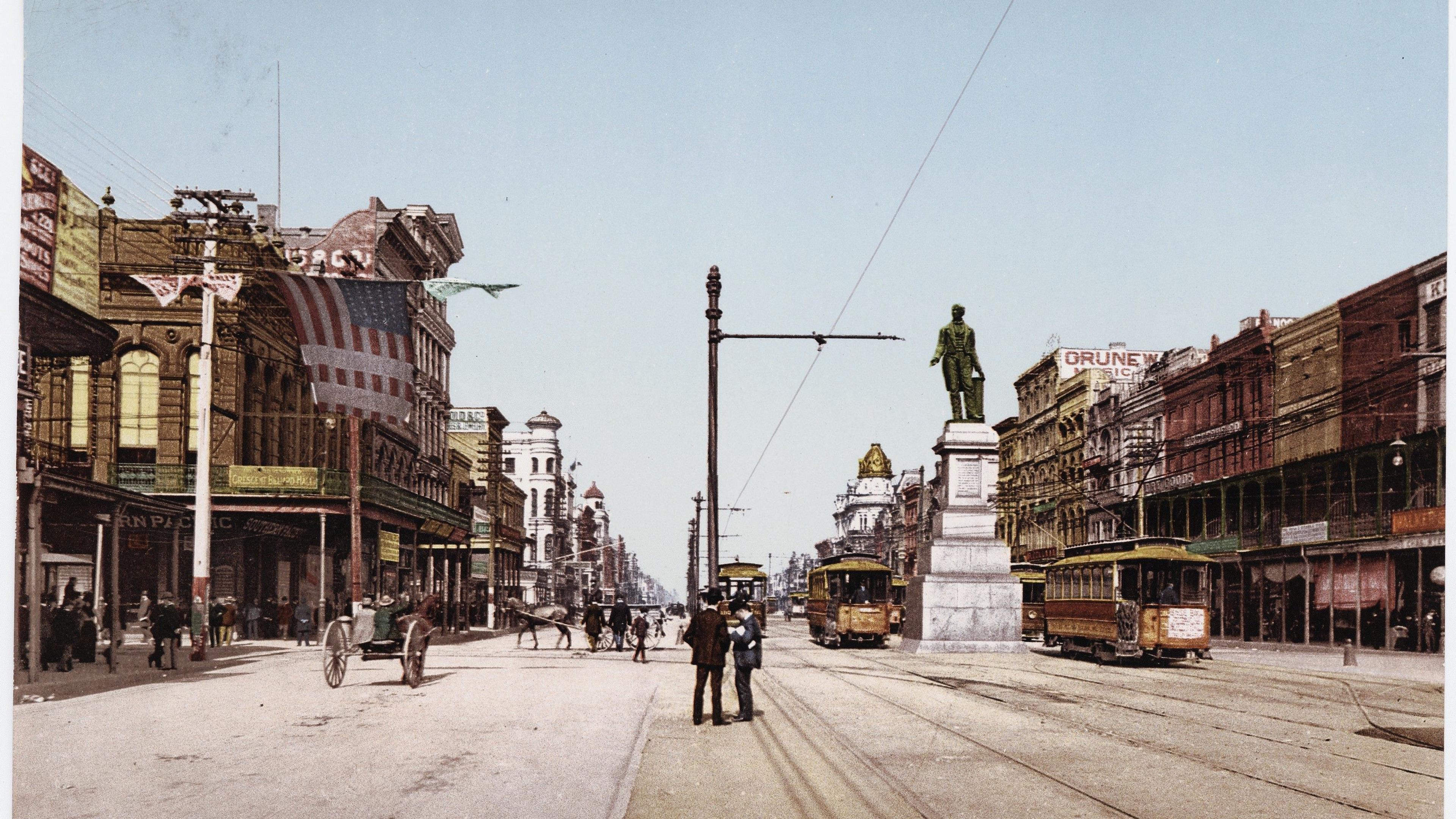Hand-coloured photo of Canal Street New Orleans featuring trams, a statue and historic buildings in early 20th century.