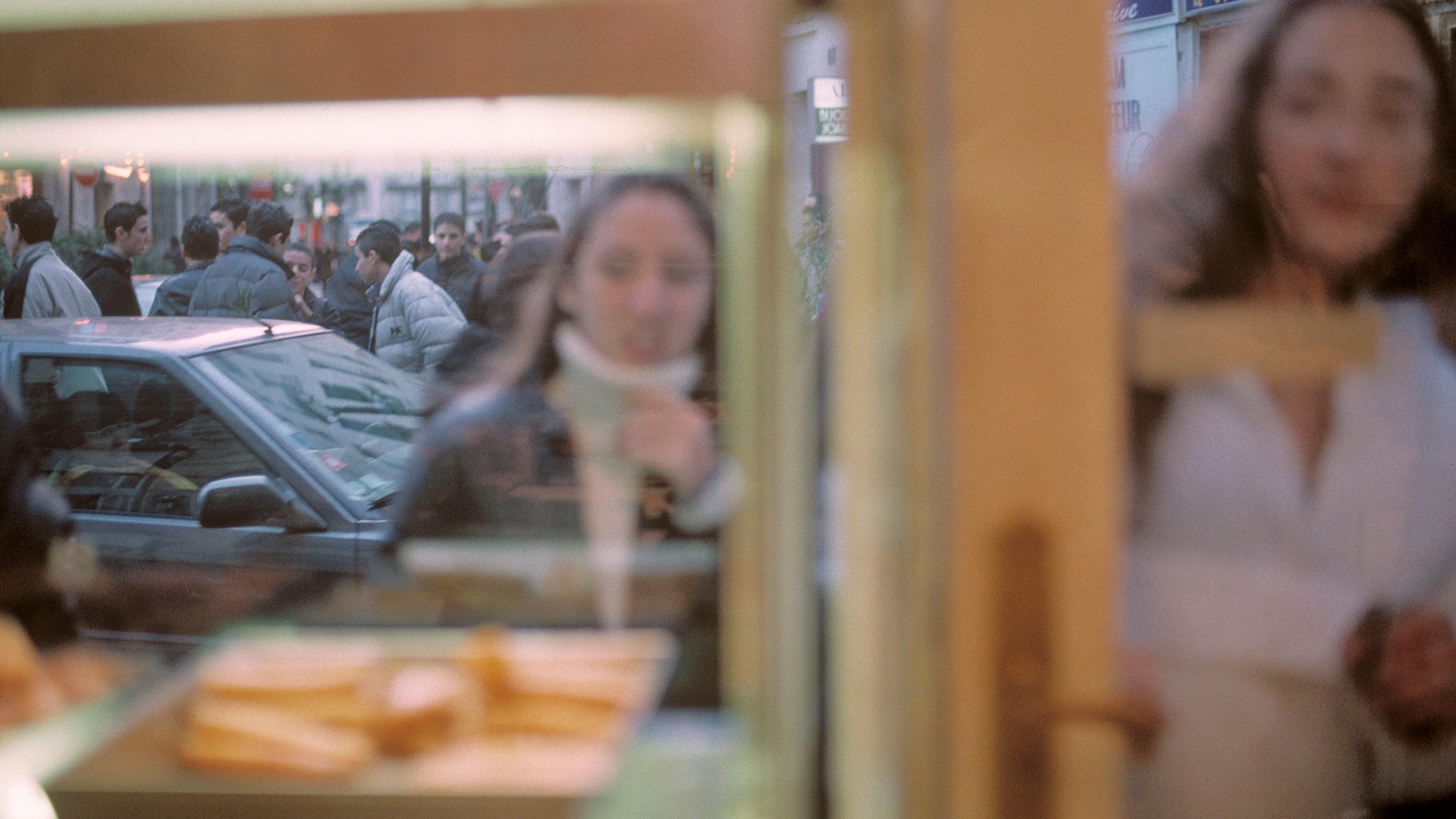Blurry photo through a shop window showing people on the street, a woman near the glass and pastry on display inside.