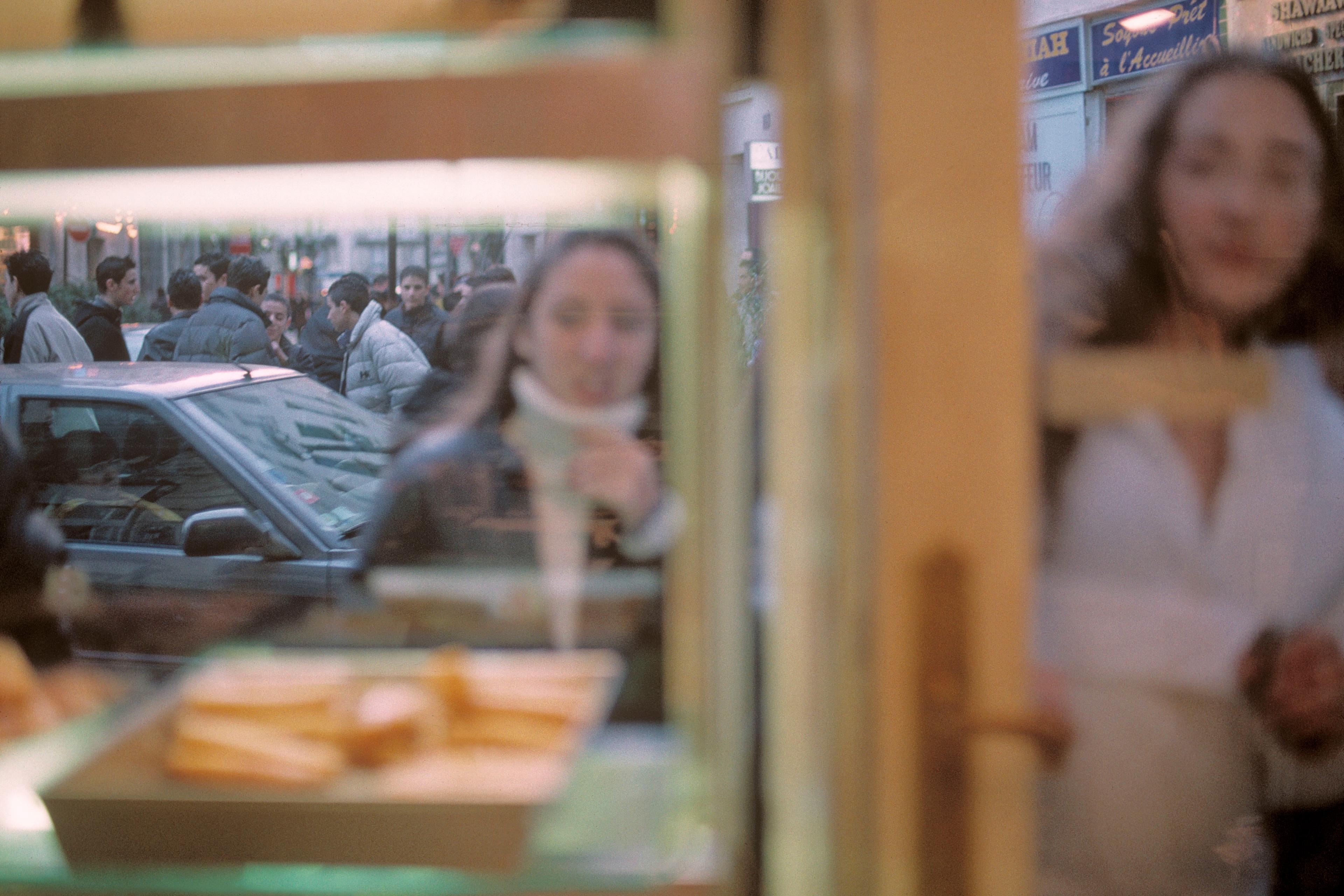 Blurry photo through a shop window showing people on the street, a woman near the glass and pastry on display inside.