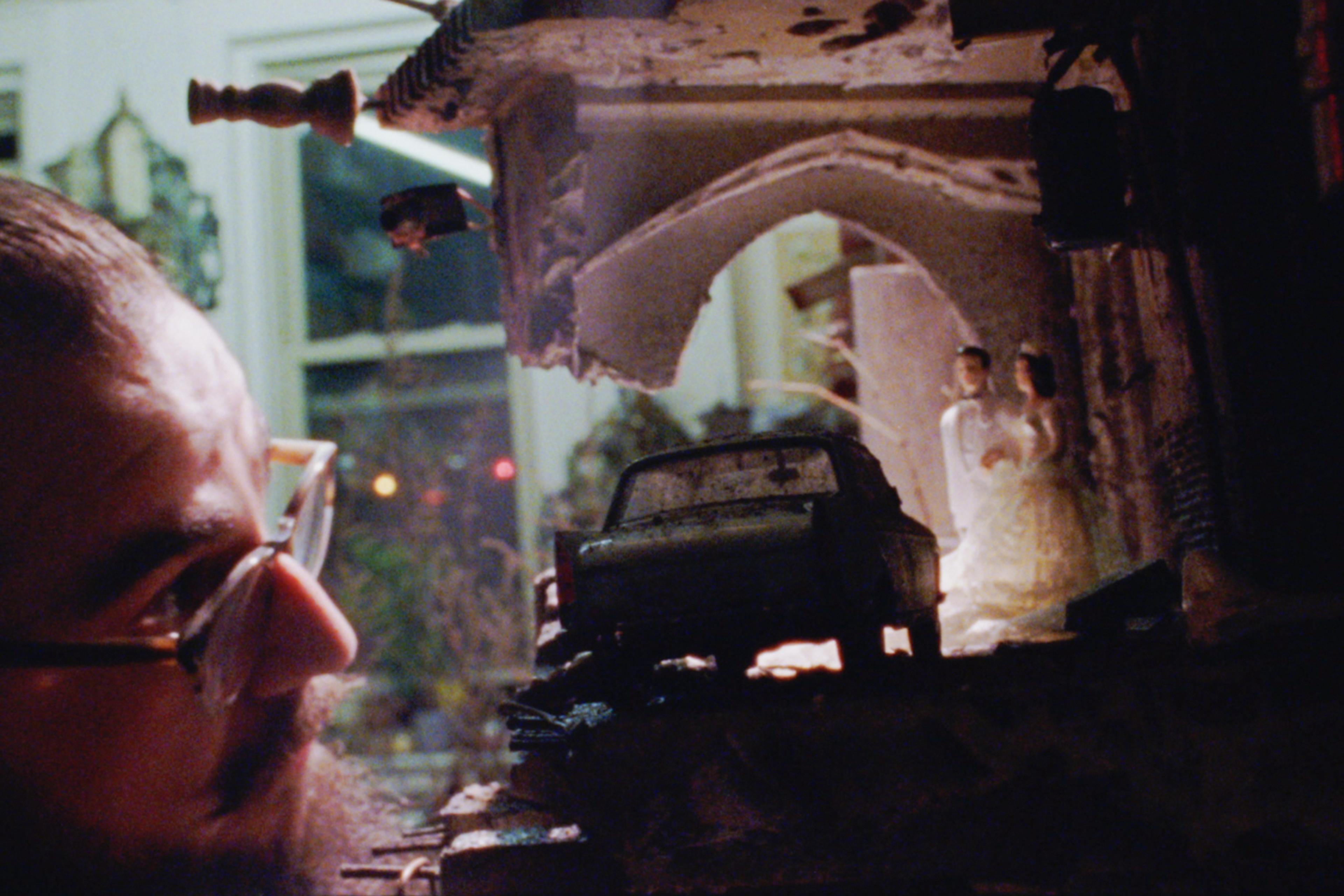 A man with glasses inspects a detailed model featuring a car, a bridge, and two figures inside a miniature building.