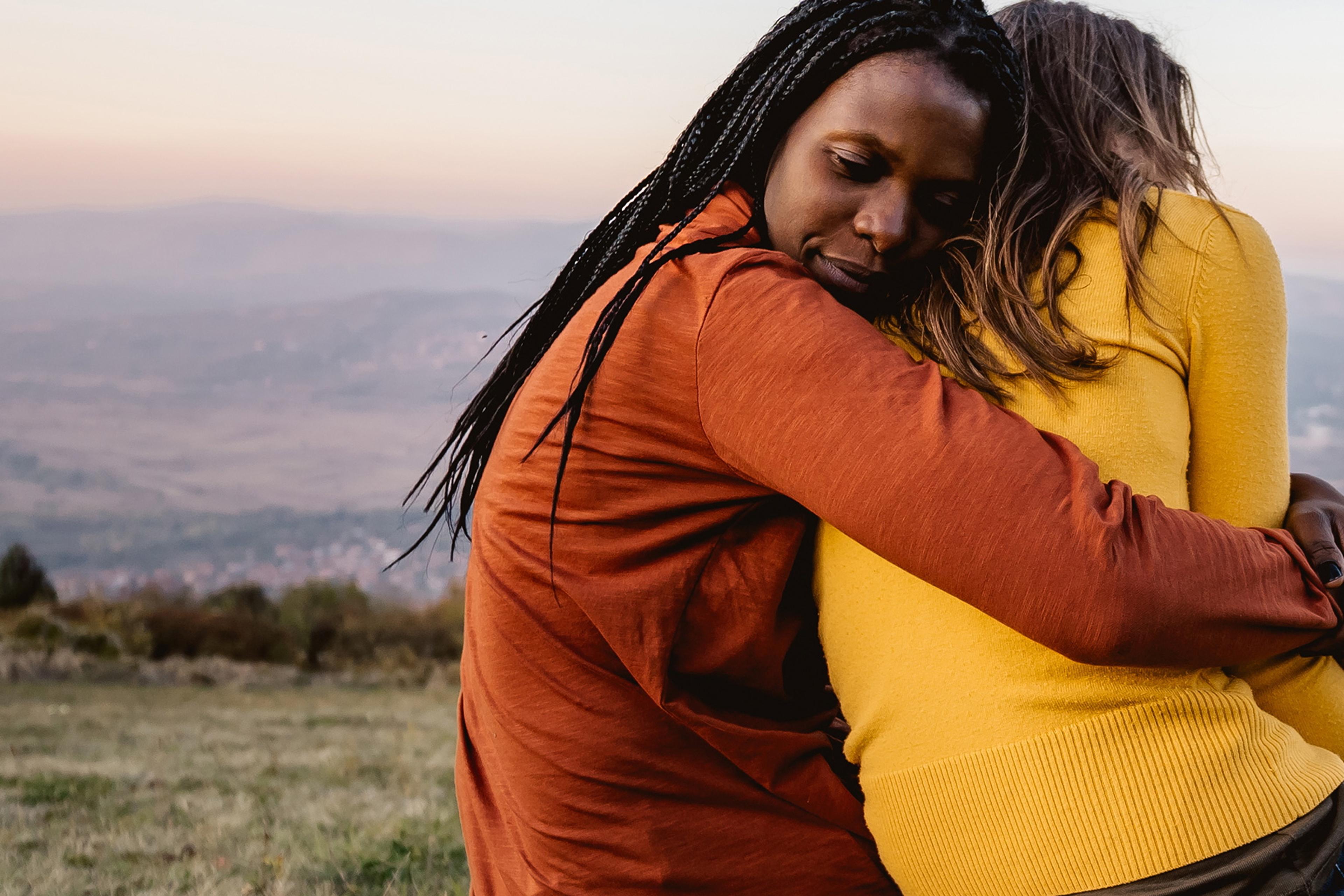 Photo of two people hugging on a grassy field with a scenic backdrop. The person on the left wears an orange jacket.