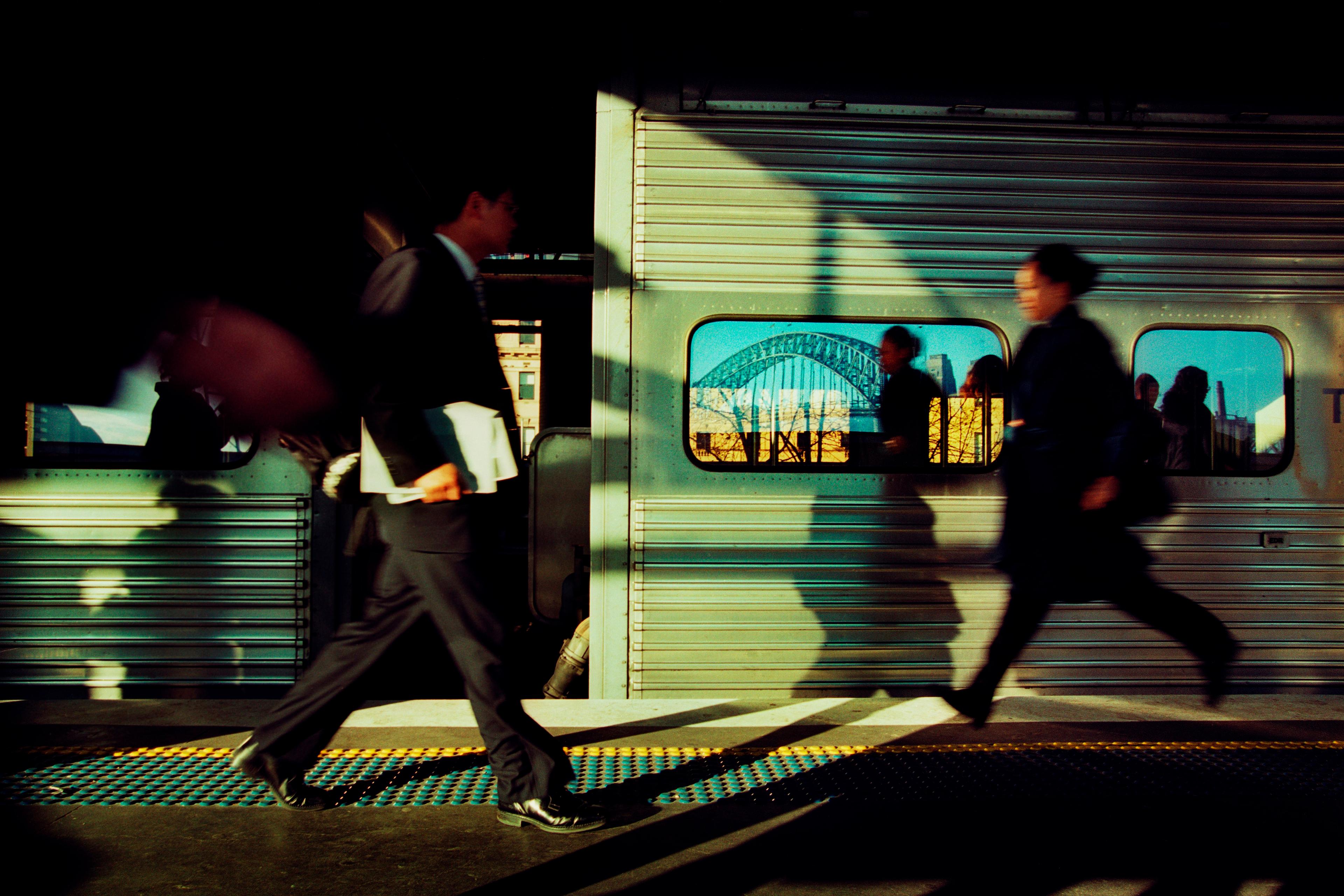 Photograph of commuters in motion at a train station with a reflection of a bridge seen in the train window.
