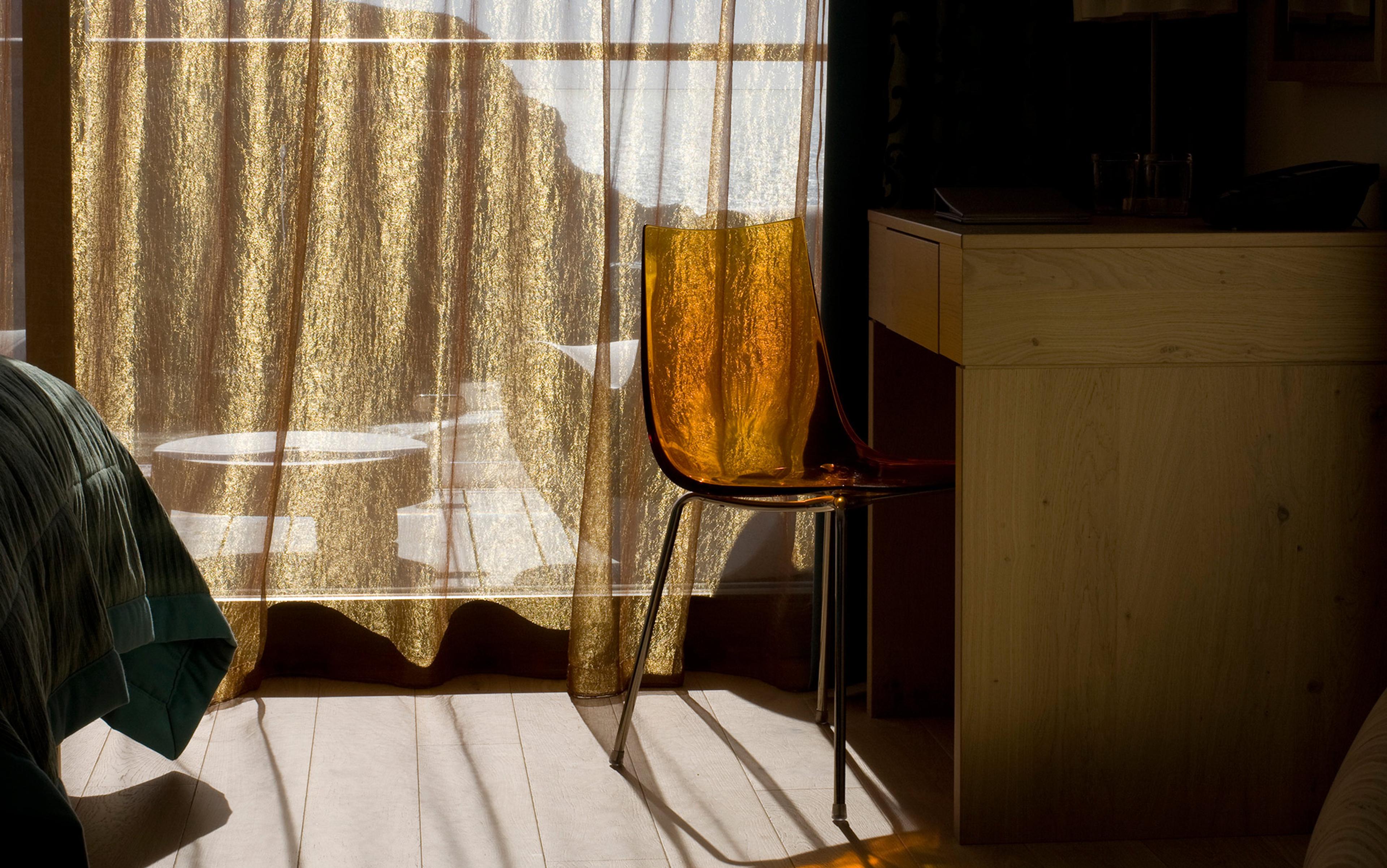A dimly lit room with sheer golden curtains, an orange transparent chair by a wooden desk, and a bed partially visible.