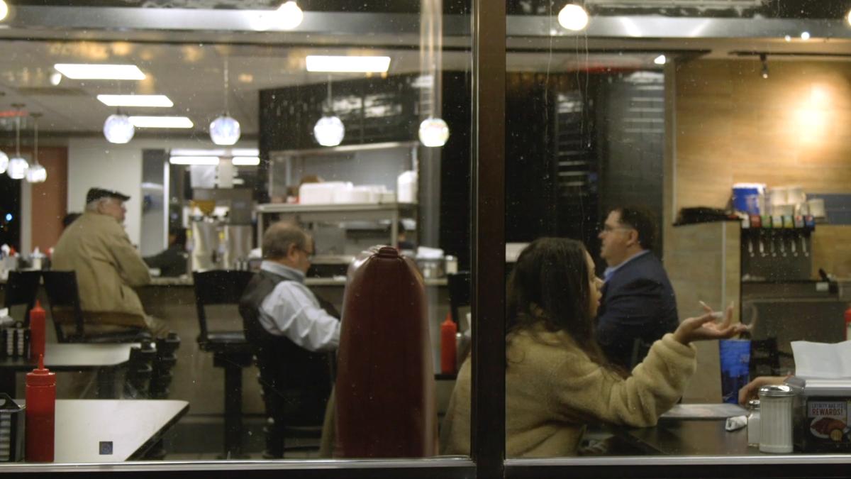 Photo of a diner interior seen through a window with patrons seated at tables and a counter under hanging lights.