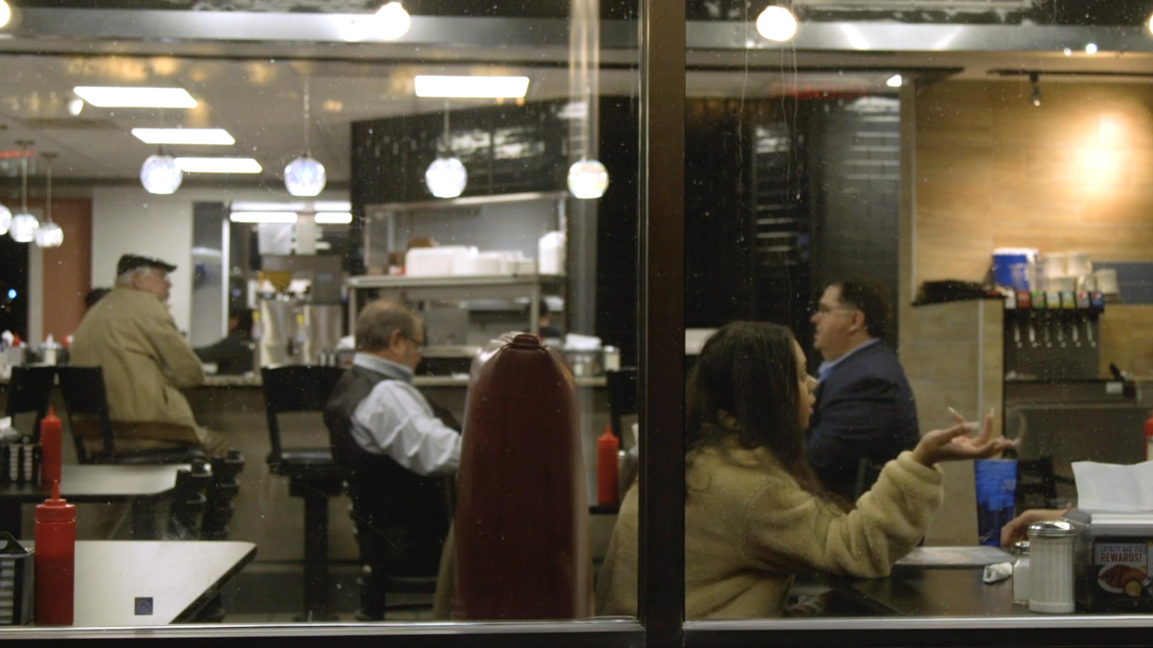 Photo of a diner interior seen through a window with patrons seated at tables and a counter under hanging lights.