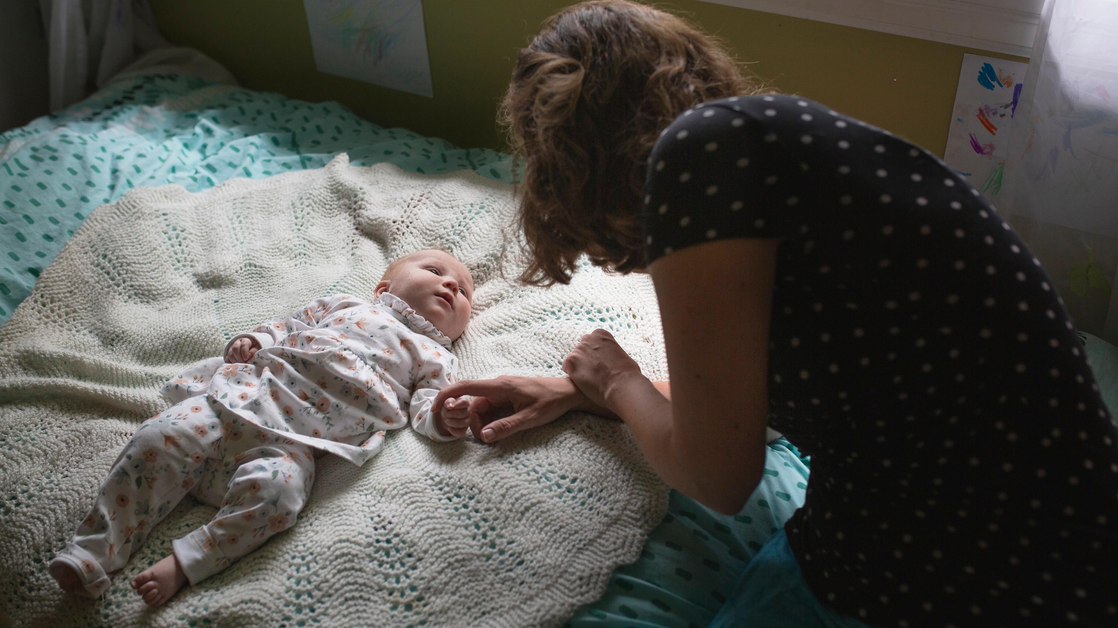Photo of a woman leaning over a baby lying on a bed, holding the baby’s hand in a softly lit room.