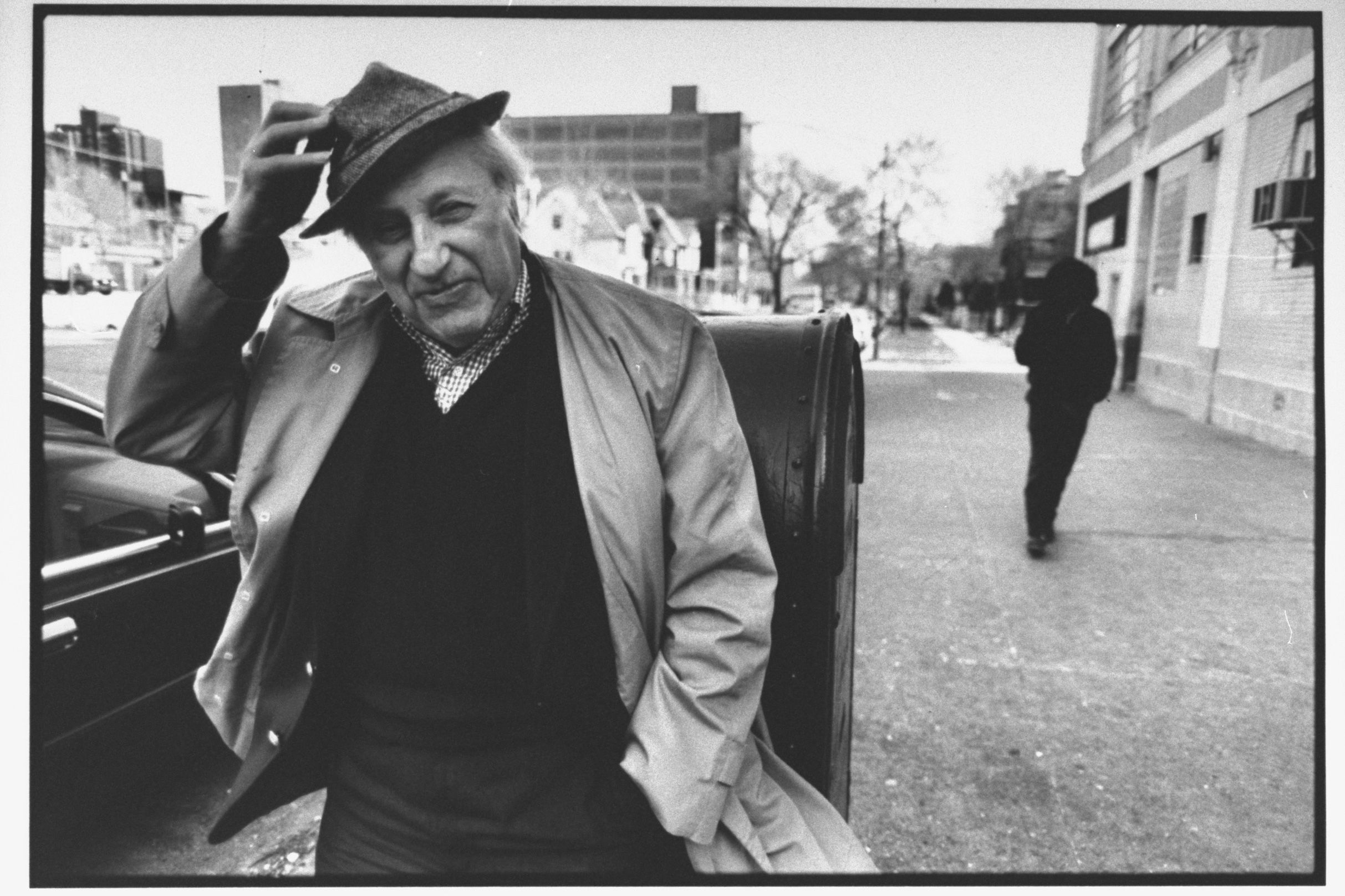 Black and white photo of an older man in a coat tipping his hat on a city street, with another person walking away.