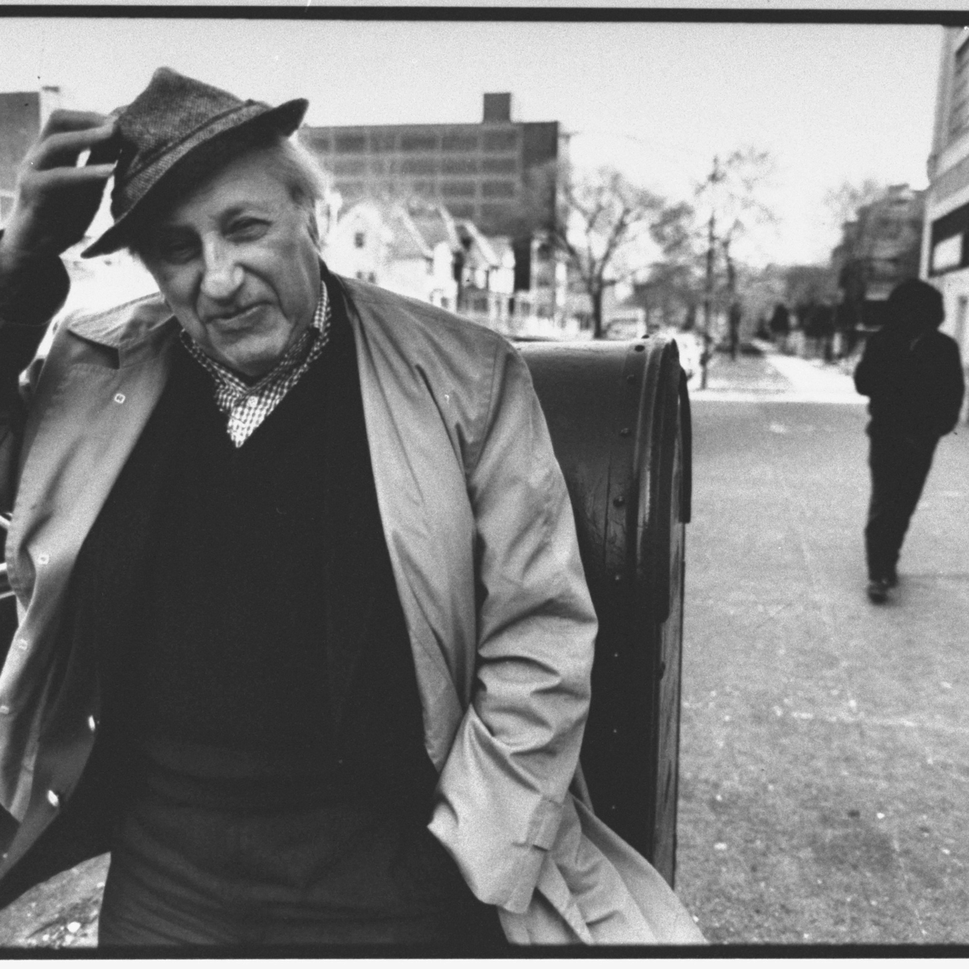 Black and white photo of an older man in a coat tipping his hat on a city street, with another person walking away.