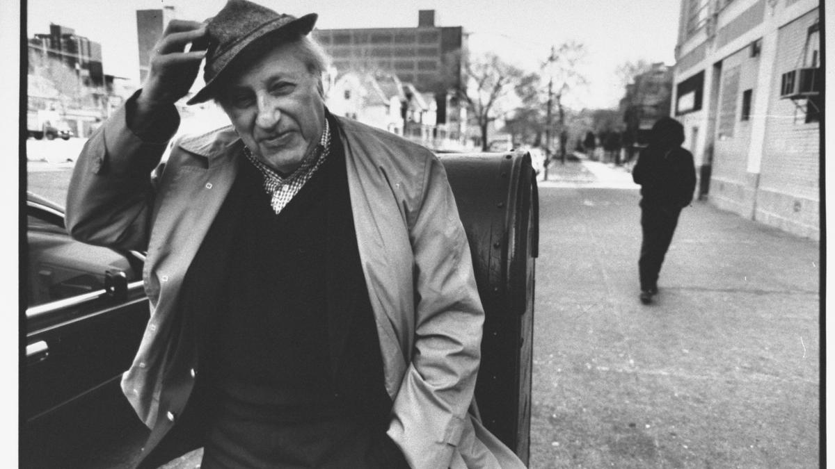 Black and white photo of an older man in a coat tipping his hat on a city street, with another person walking away.