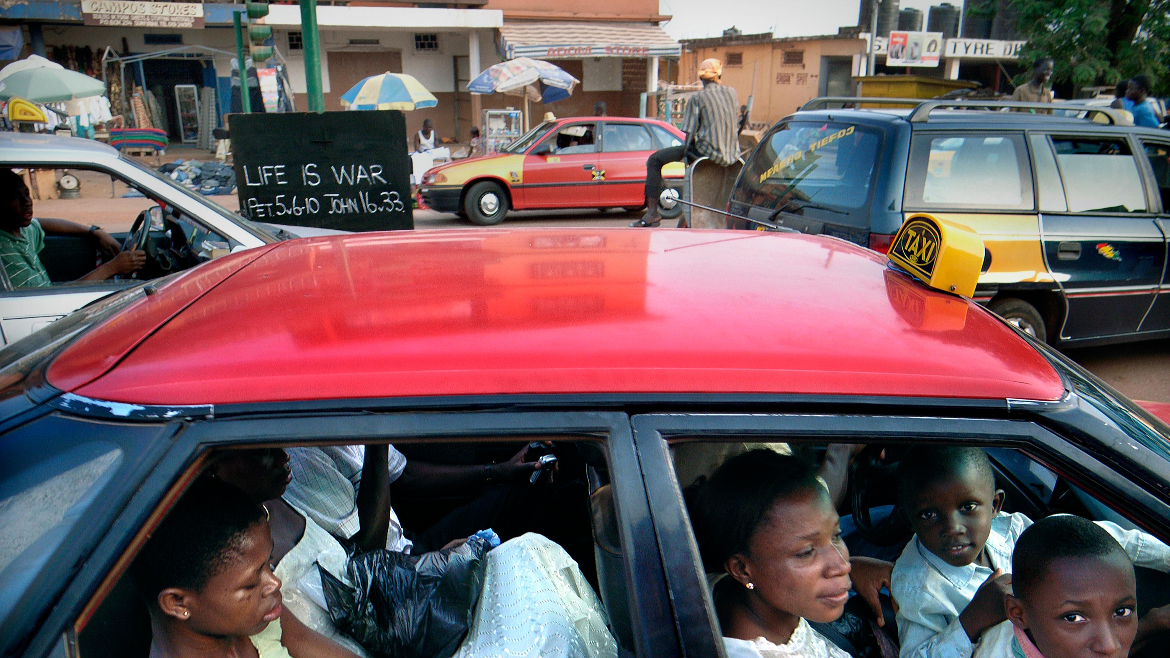 Photo of a red taxi with passengers in an urban setting, a sign saying “Life is War” visible in the background.