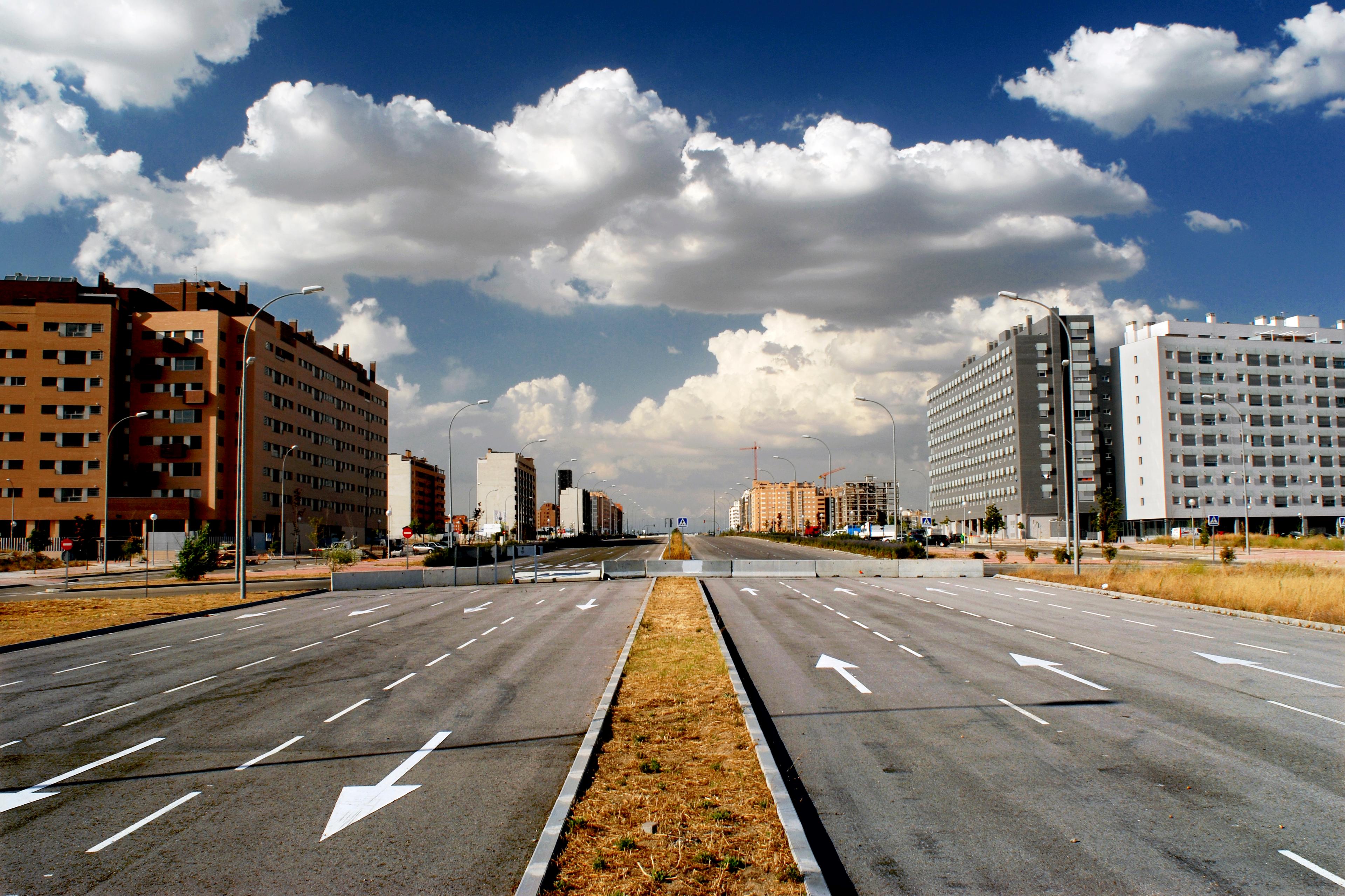 A wide empty road with buildings on either side under a blue sky filled with fluffy clouds.