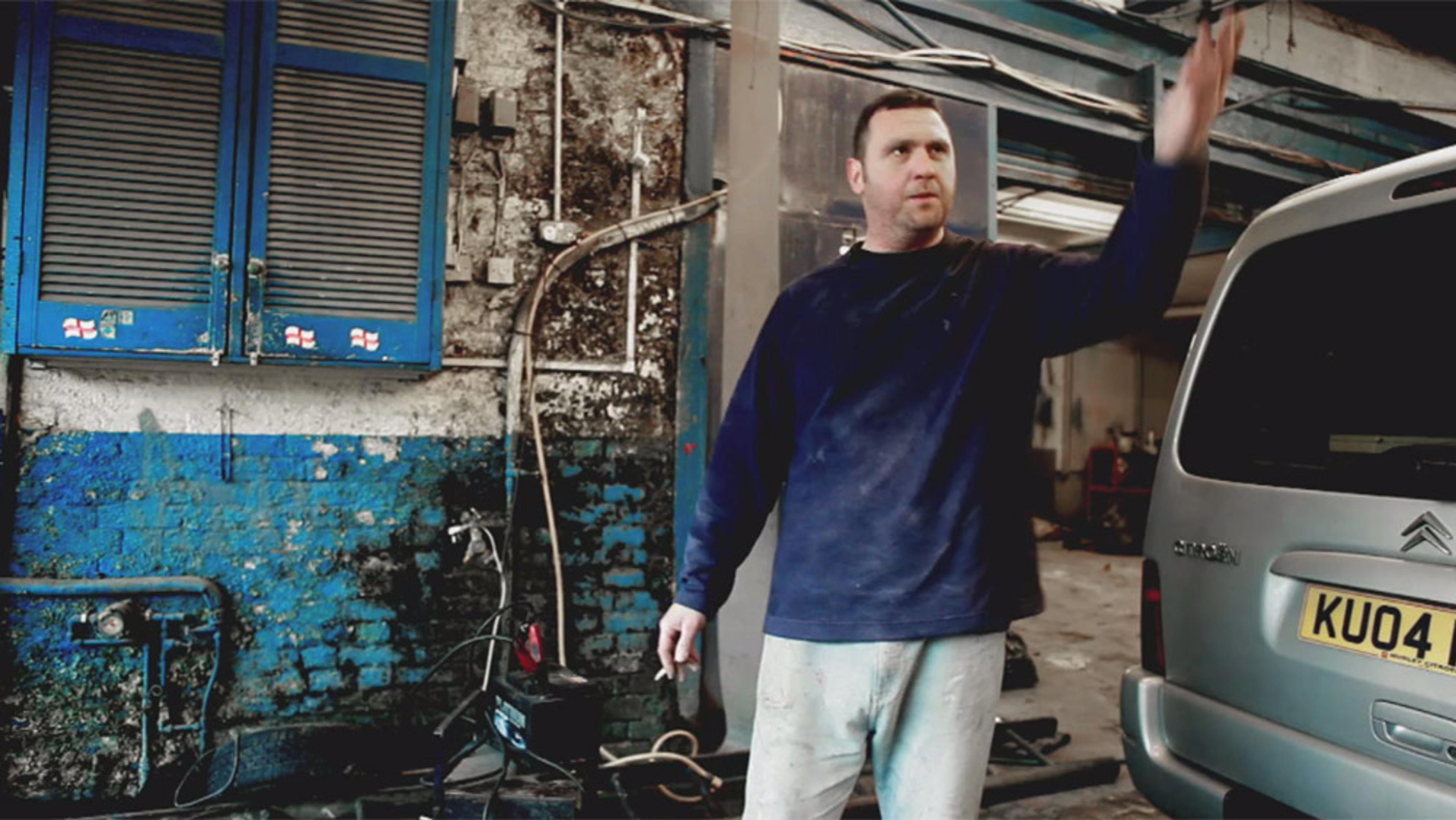 A man in a blue jumper and light trousers in a gritty garage with blue cabinets, weathered walls and a silver car.