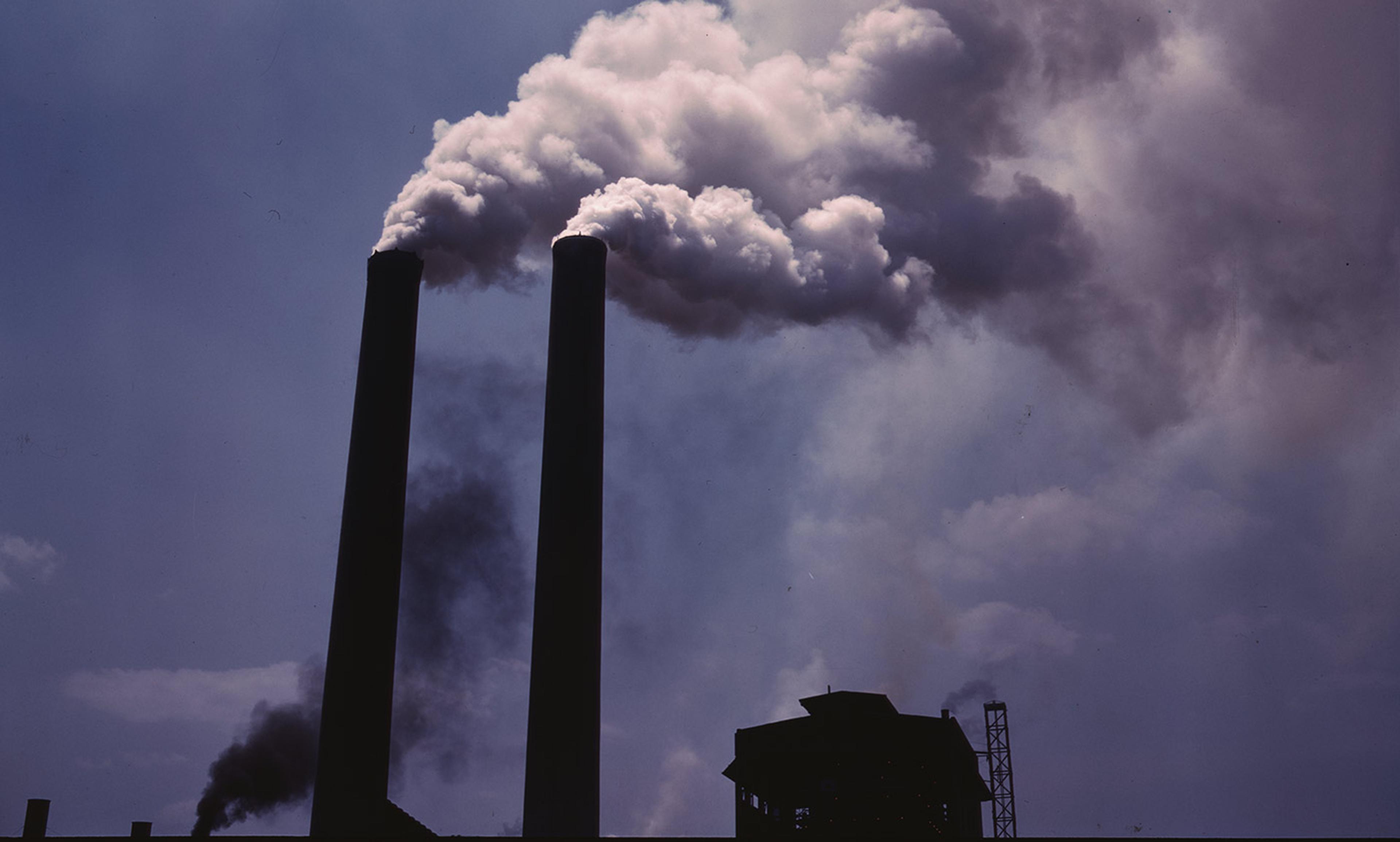 Two large smokestacks emitting thick clouds of smoke against a dark sky with an industrial structure silhouetted below.