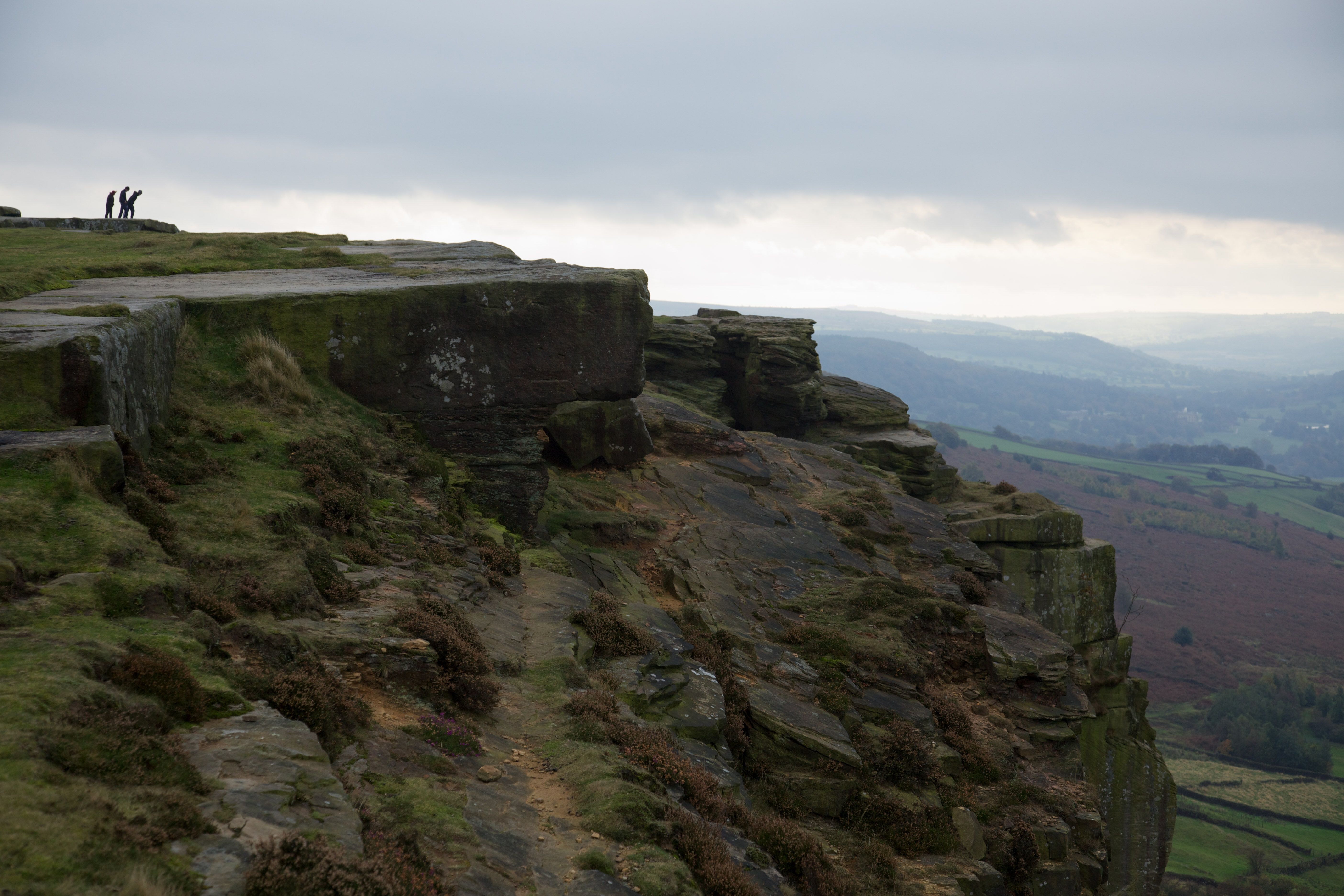 Photo of a rugged cliff edge with two people in the distance, overlooking a misty landscape under a cloudy sky.