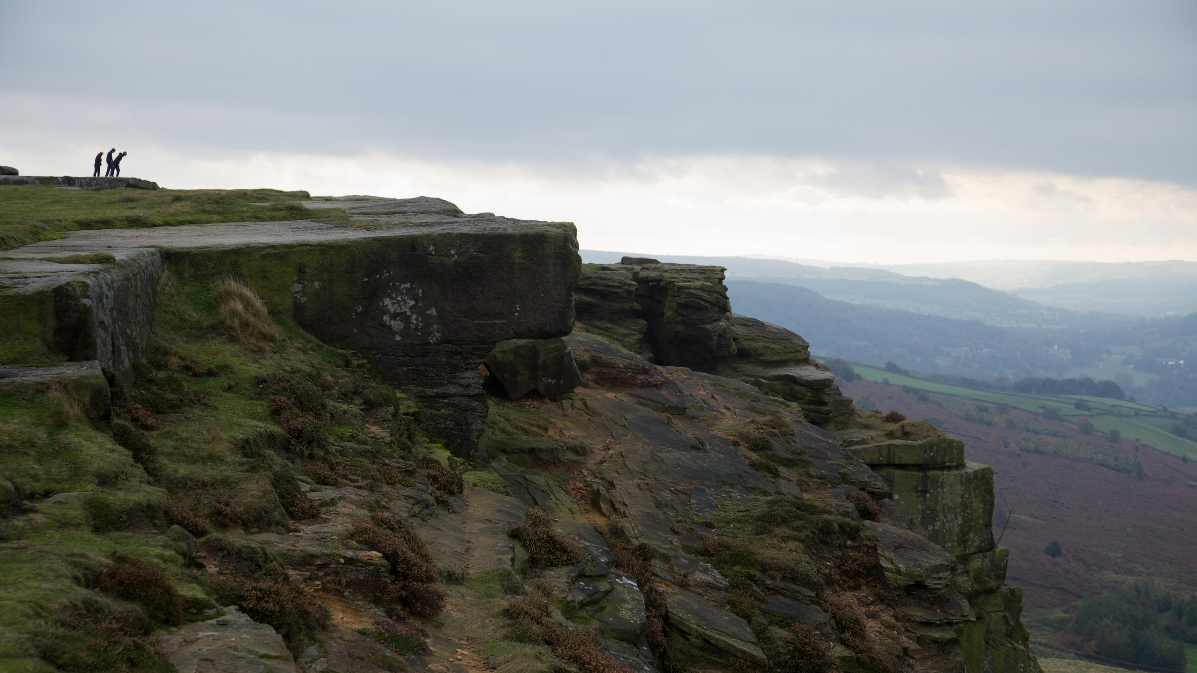 Photo of a rugged cliff edge with two people in the distance, overlooking a misty landscape under a cloudy sky.