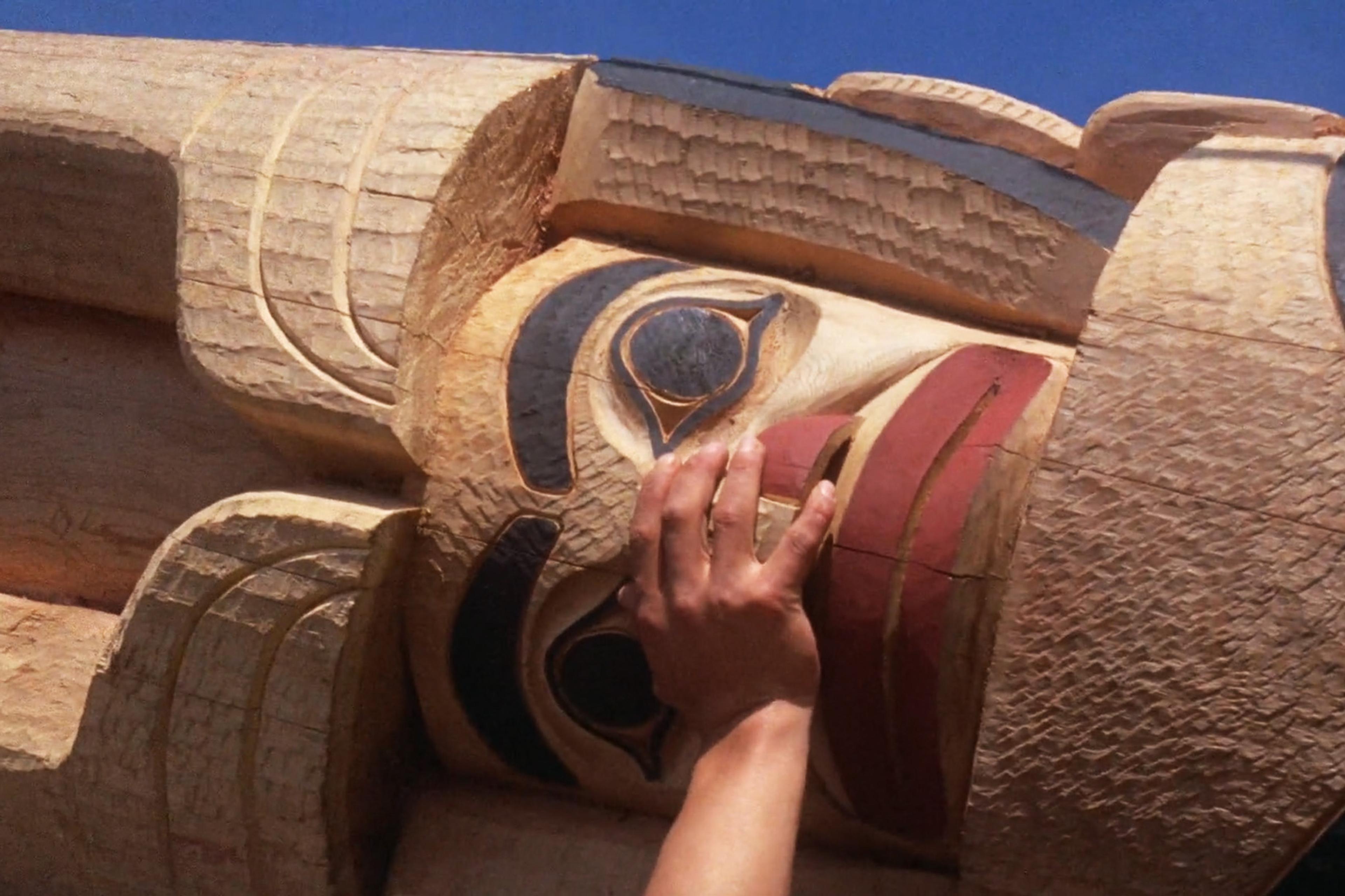 Hand touching a detailed wooden totem pole carving with traditional painted designs; clear blue sky in the background.