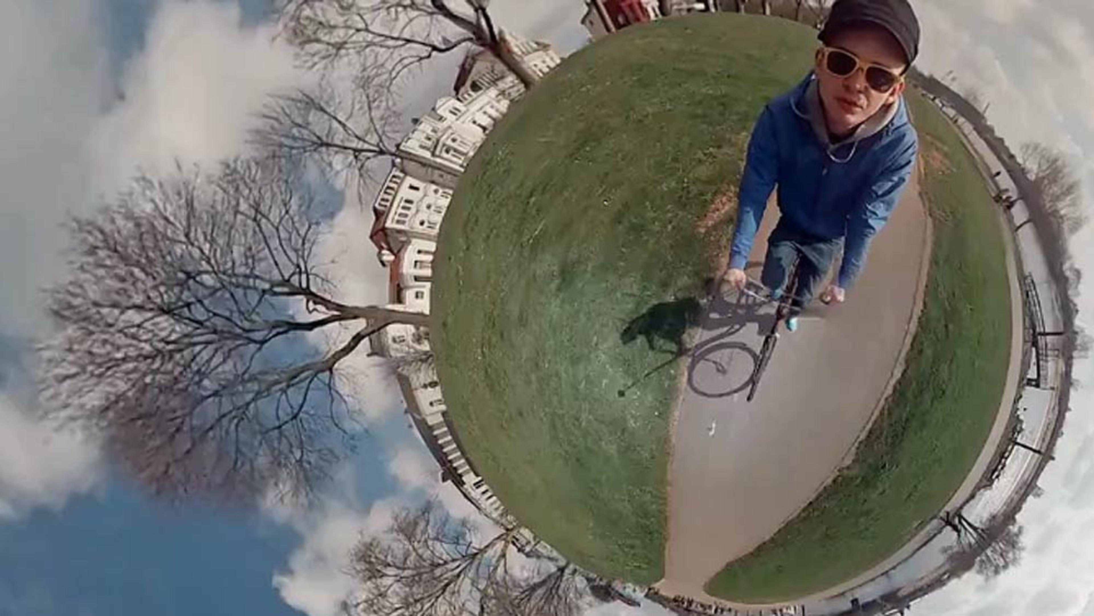 Fisheye photo of a man cycling on a path wearing sunglasses and a cap with houses, trees, and sky distorted around him.