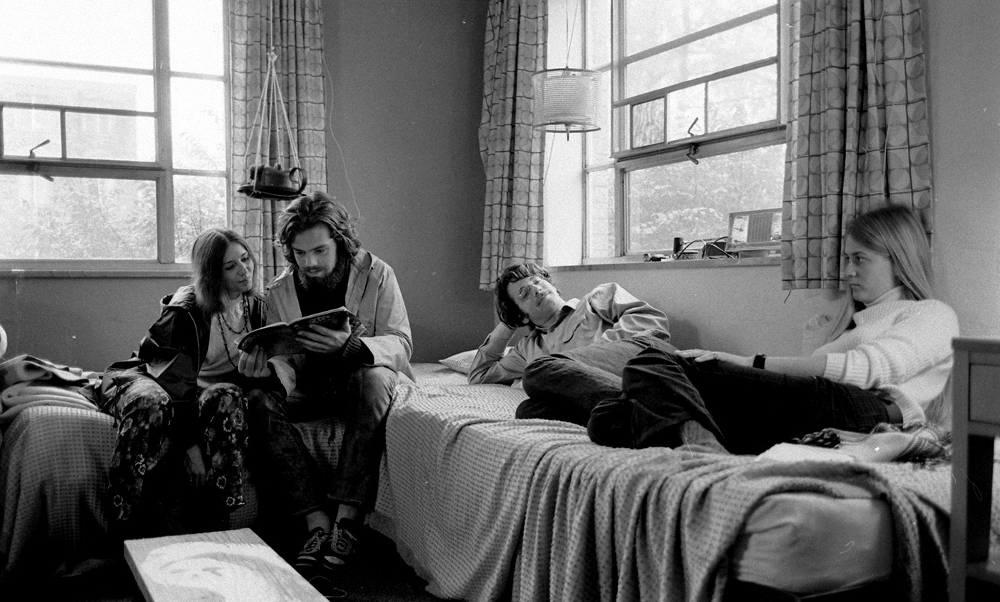 A black-and-white photo of four people in a room: a couple looking at a book, a man lying on a bed looking happily at a woman who sits gloomy nearby.