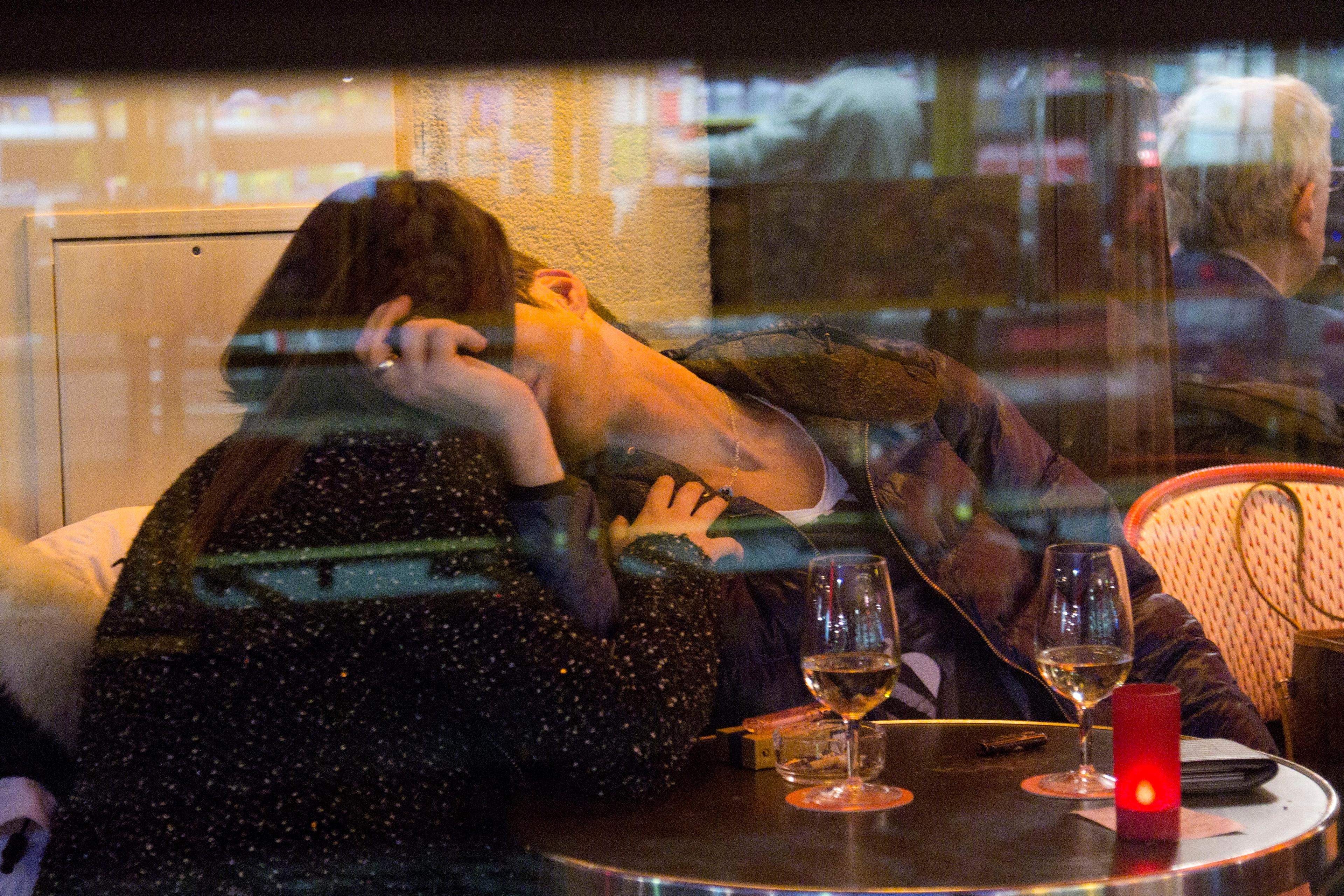 Photo of a couple kissing at a café table with glasses of wine, seen through a window reflecting city lights.