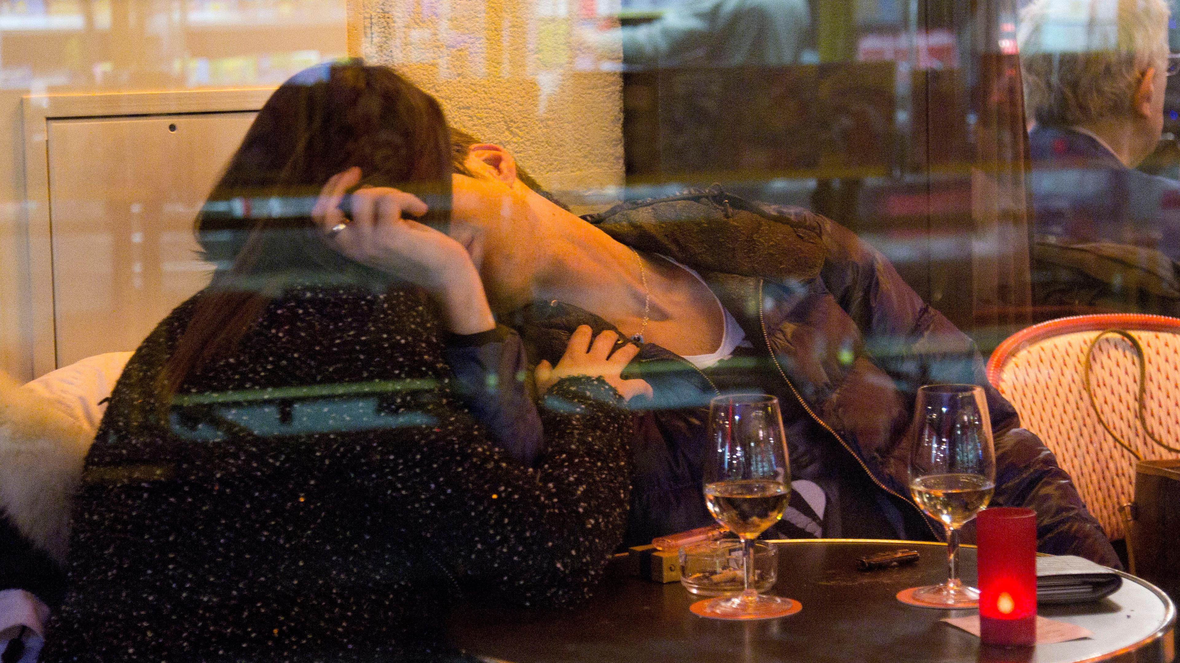 Photo of a couple kissing at a café table with glasses of wine, seen through a window reflecting city lights.
