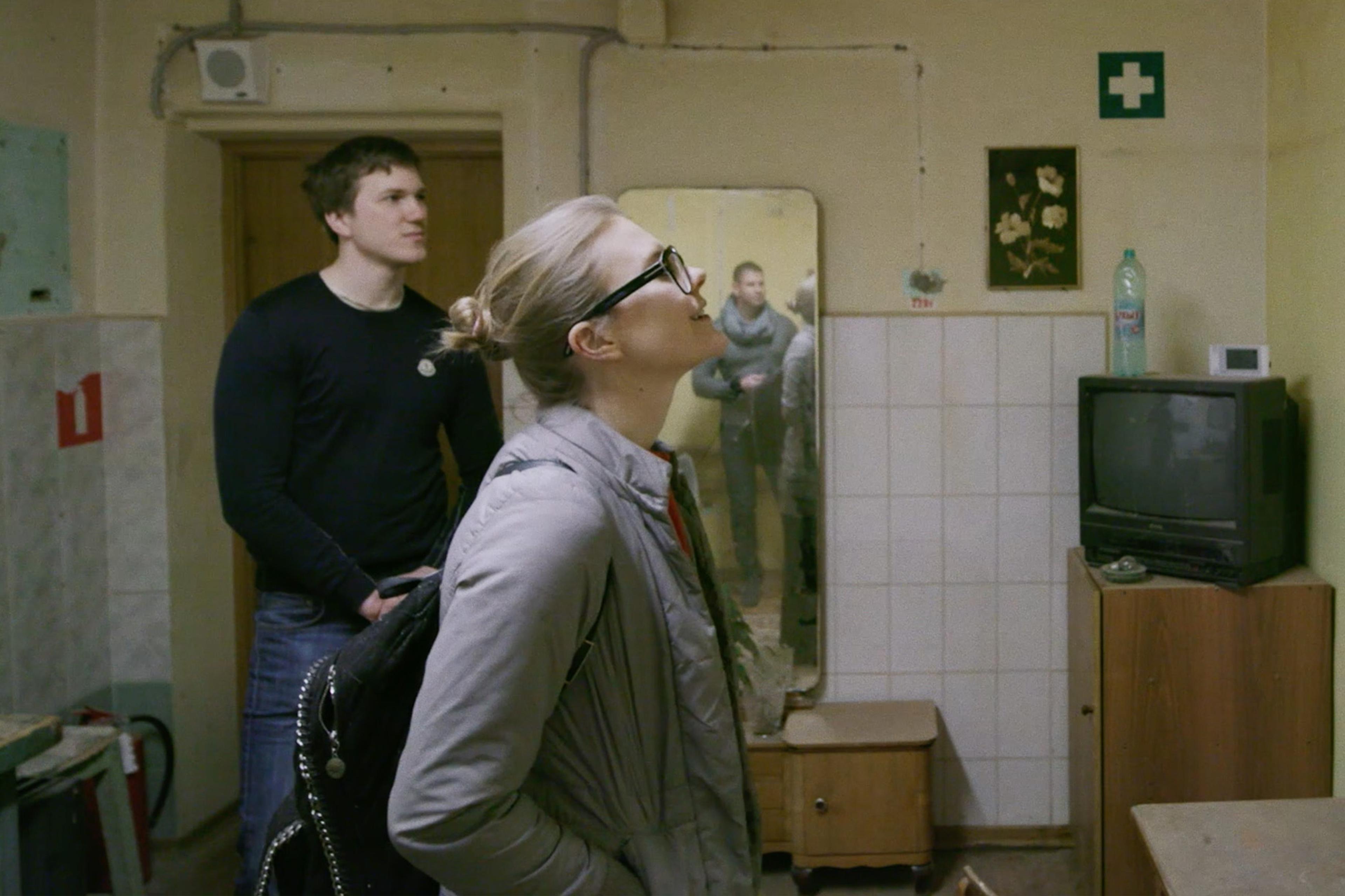 Two people in a worn-out room with an old TV, a mirror reflecting more people, and various items on countertops and shelves.