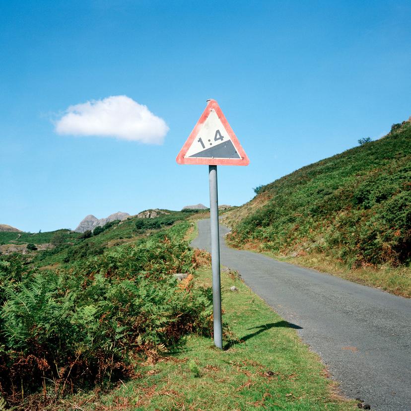 A steep road with a 1:4 gradient warning sign, flanked by greenery and hills under a clear blue sky.