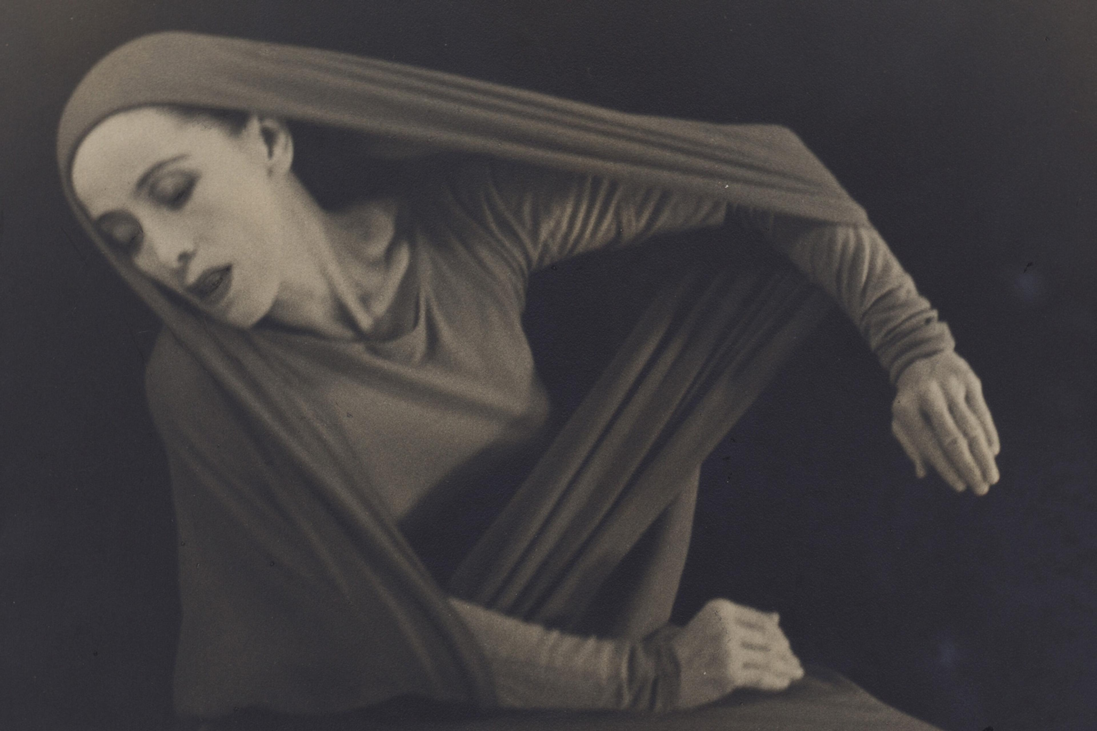 A black-and-white photo of a dancer in a flowing, long-sleeved outfit, posed with eyes closed and hands extended.