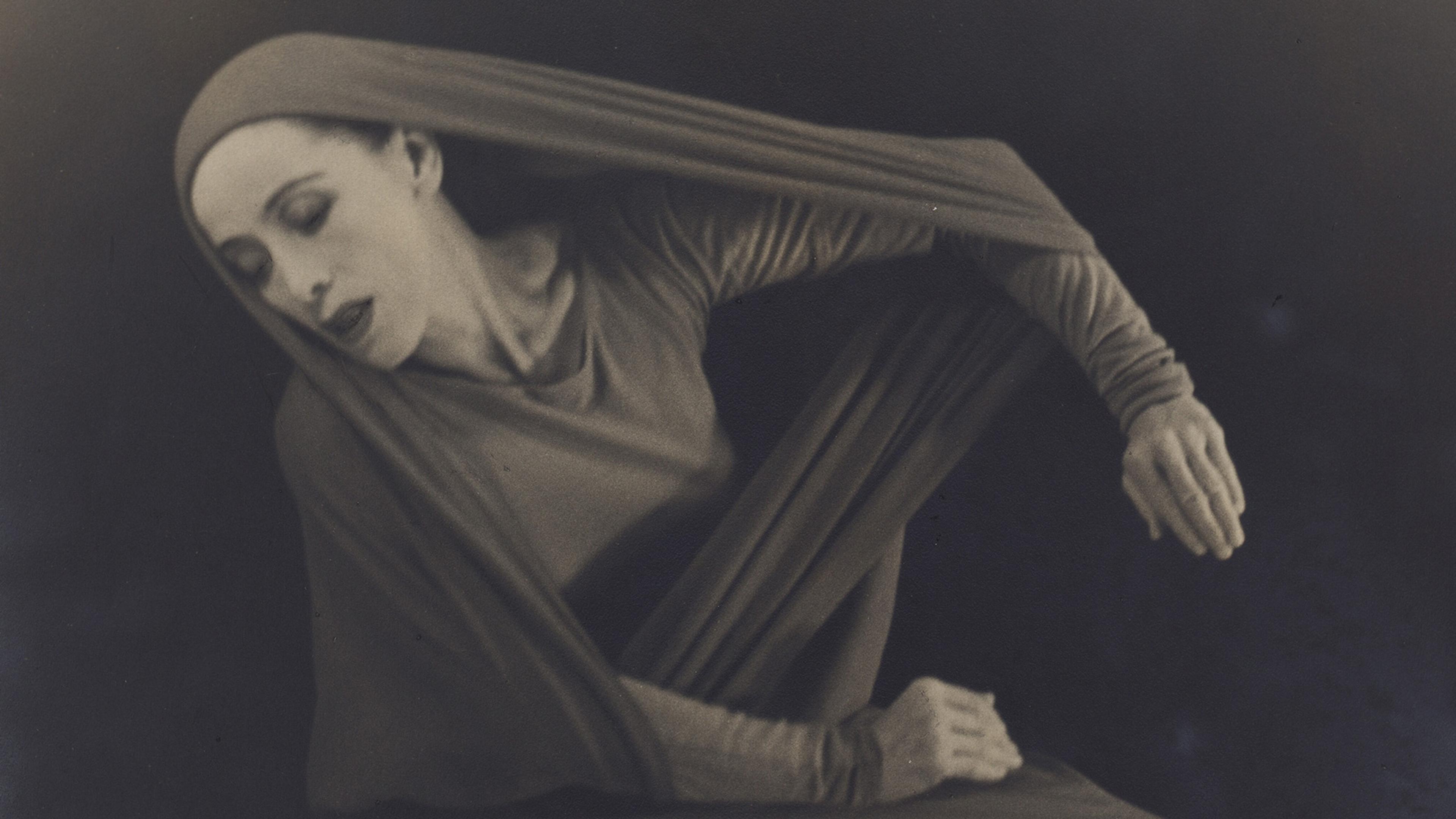 A black-and-white photo of a dancer in a flowing, long-sleeved outfit, posed with eyes closed and hands extended.