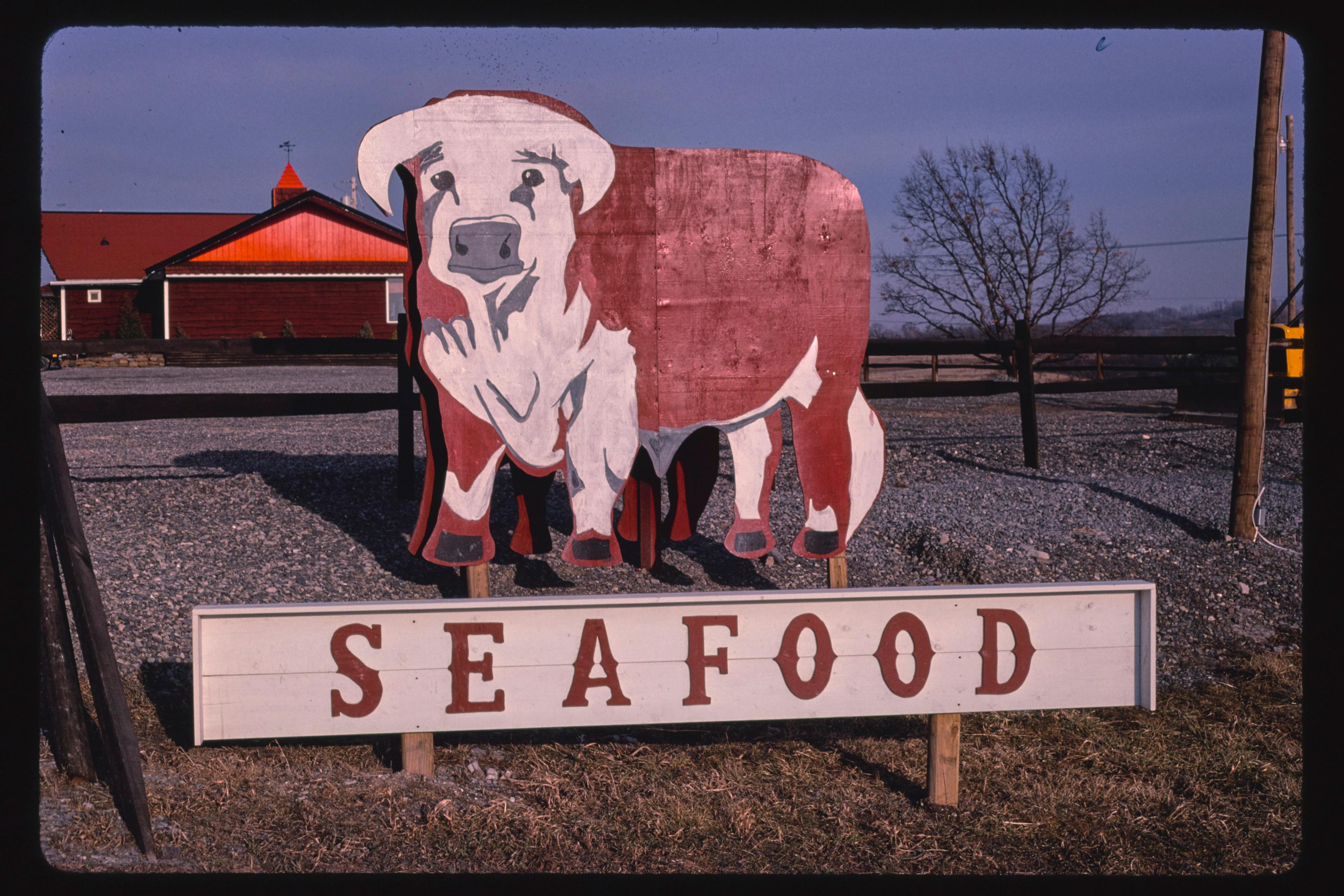 Photo of a red and white cow sign above the word “SEAFOOD” against a rural backdrop with a red barn.
