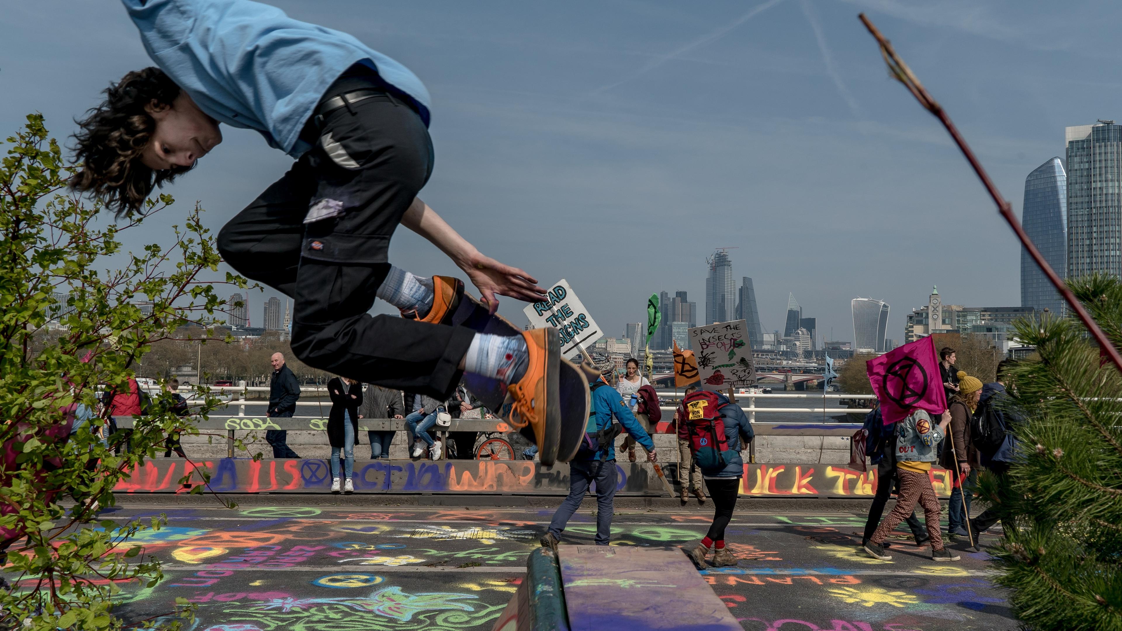 Photo of a person jumping over graffiti-covered pavement with protesters in the background holding environmental signs.