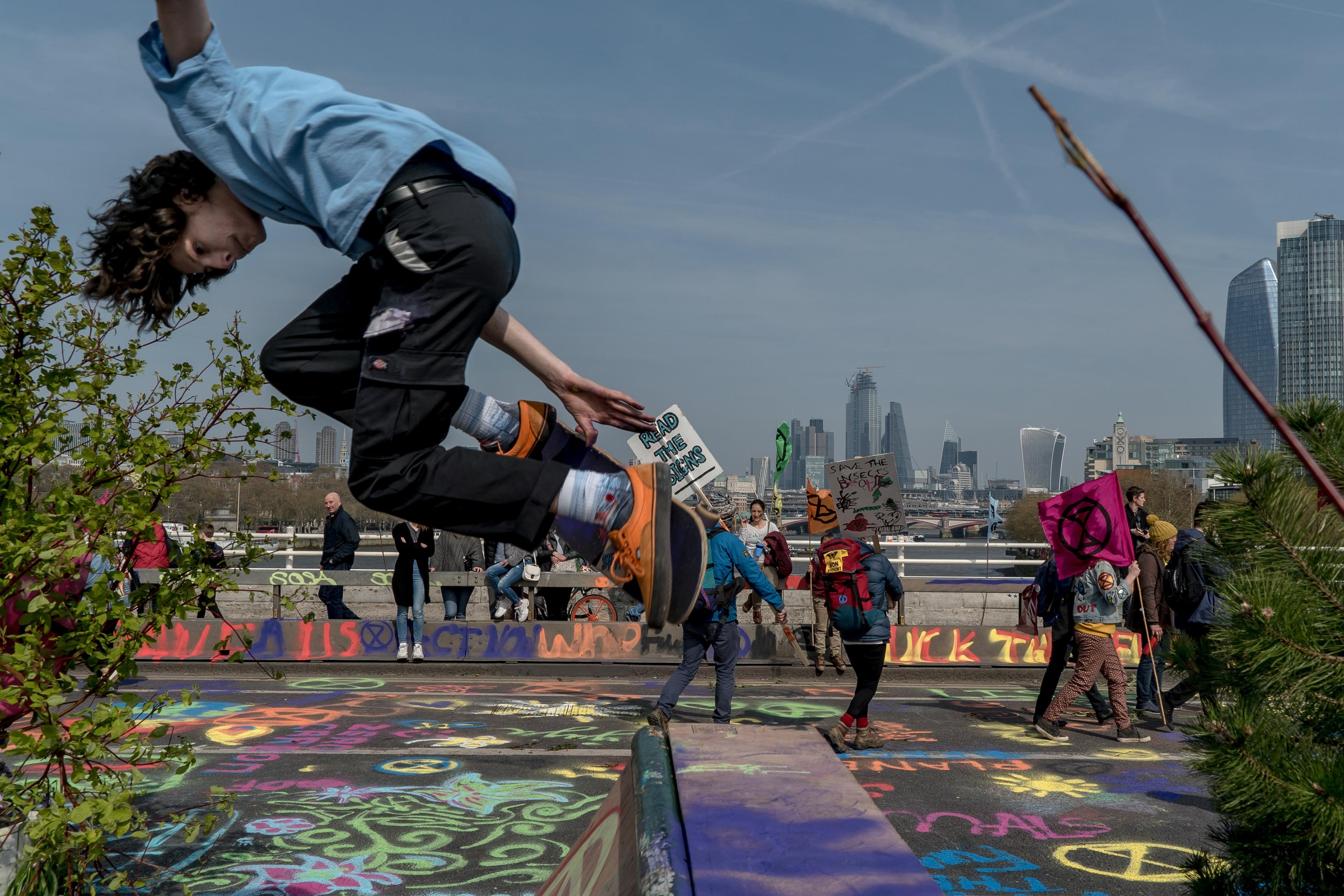 Photo of a person jumping over graffiti-covered pavement with protesters in the background holding environmental signs.