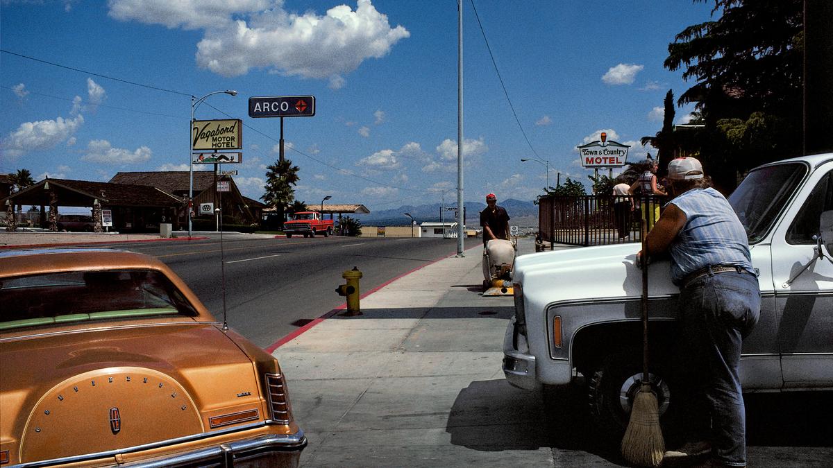 Photo of a street with vintage cars, motel signs and a person leaning on a truck holding a broom under a blue sky.