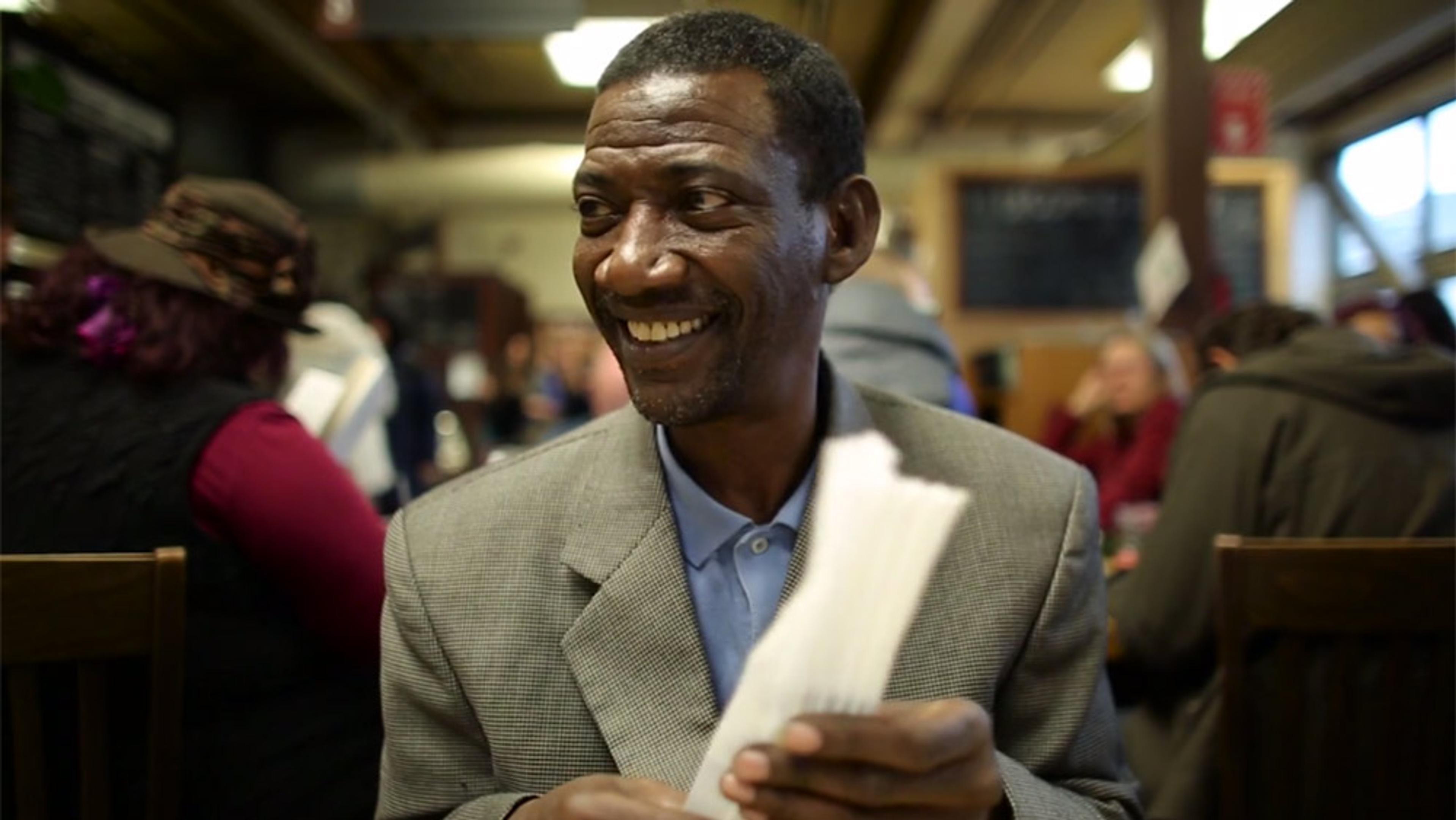 A man smiling, seated at a table in a busy indoor setting, holding a stack of papers with people in the background.