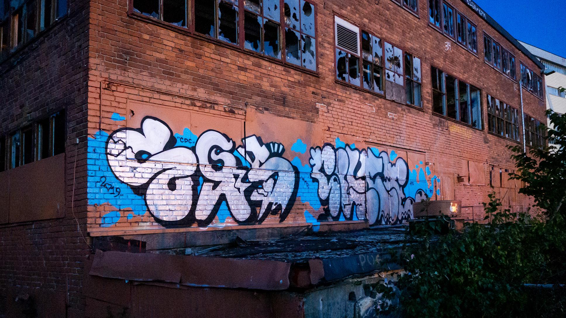 Photo of a derelict brick building with colourful graffiti on the wall broken windows and overgrown vegetation at dusk.