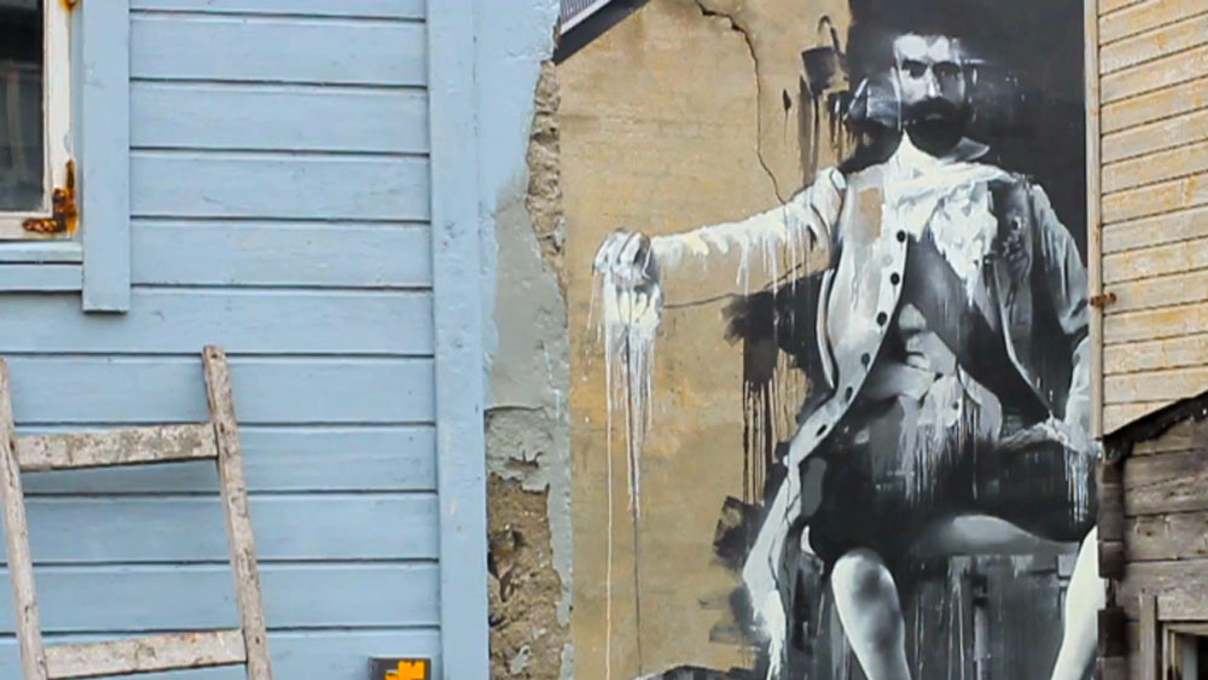 Street art of a man in a suit on a decaying wall between blue and beige buildings with a ladder leaning against the blue wall.
