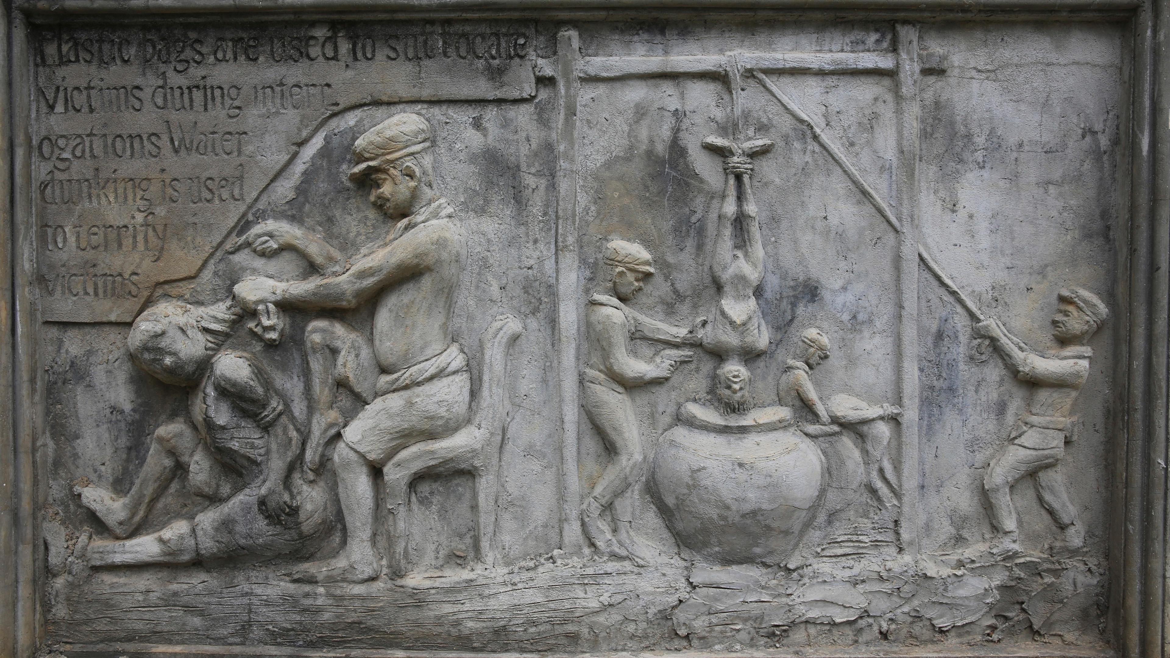 Stone relief depicting interrogation methods with figures enacting scenes of torture, accompanied by descriptive text.
