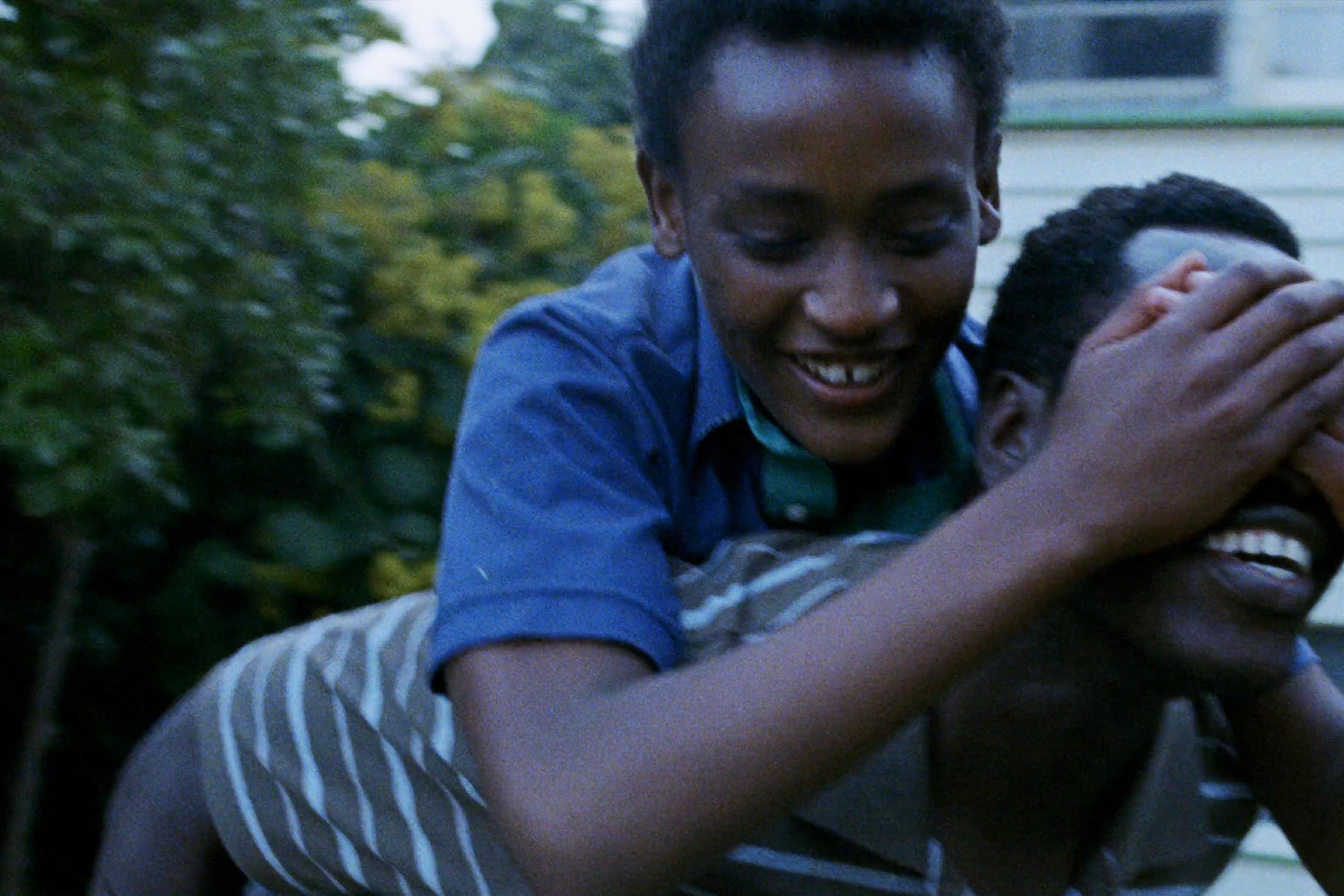 Two Black men outdoors at dusk, the older is giving the younger one a piggyback ride, both smiling and playful.