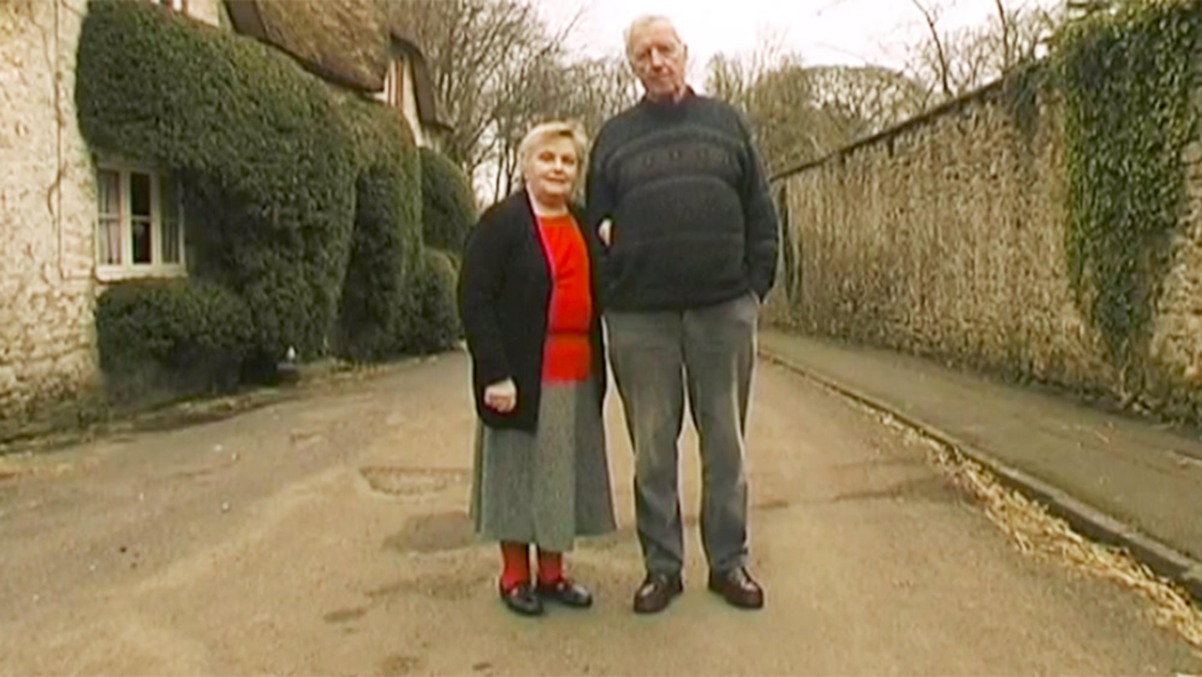 An elderly couple standing on a village street next to a stone house with a thatched roof and large hedges.