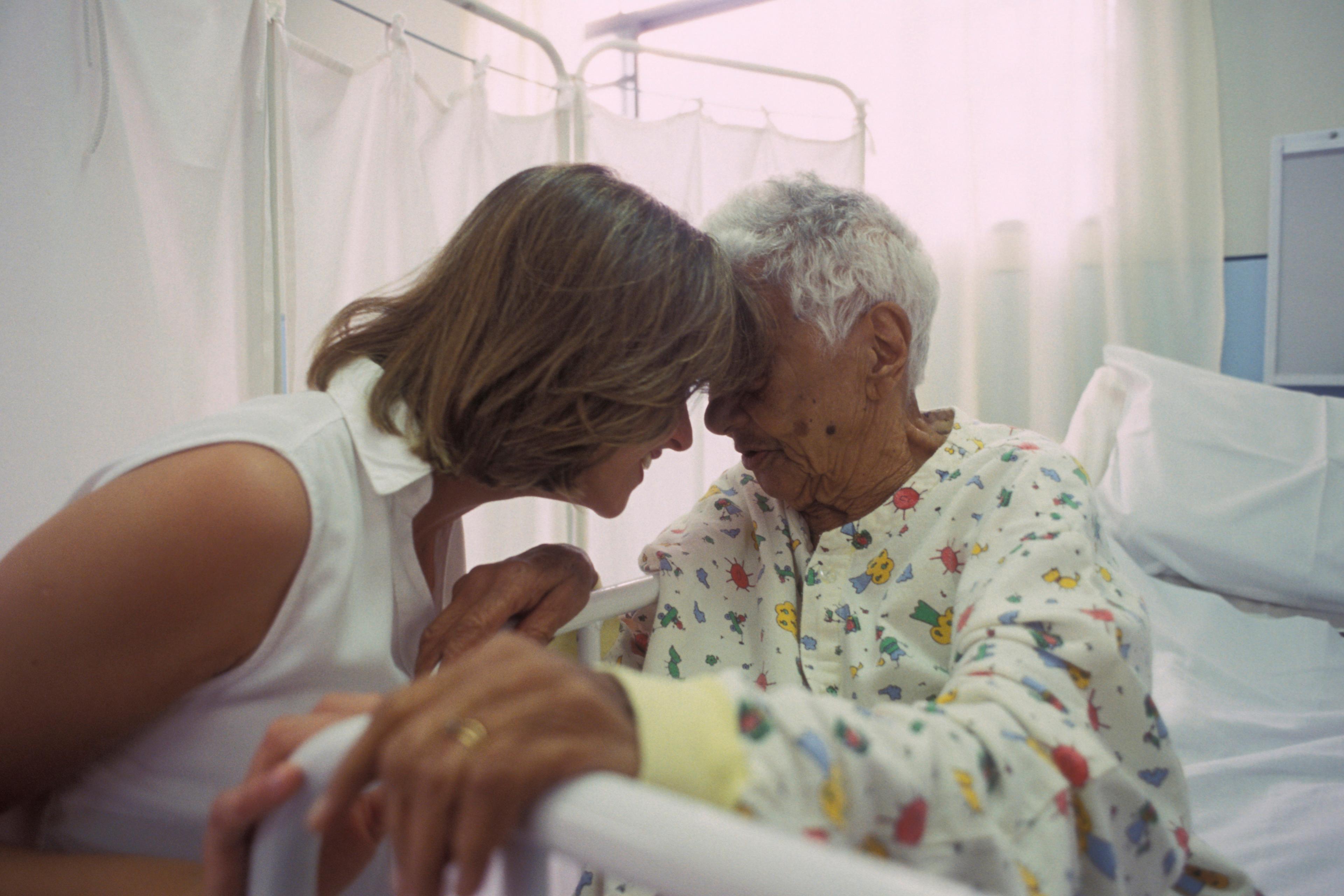 Photo of a woman in white comforting an elderly woman in a hospital gown with their foreheads touching in a hospital room.