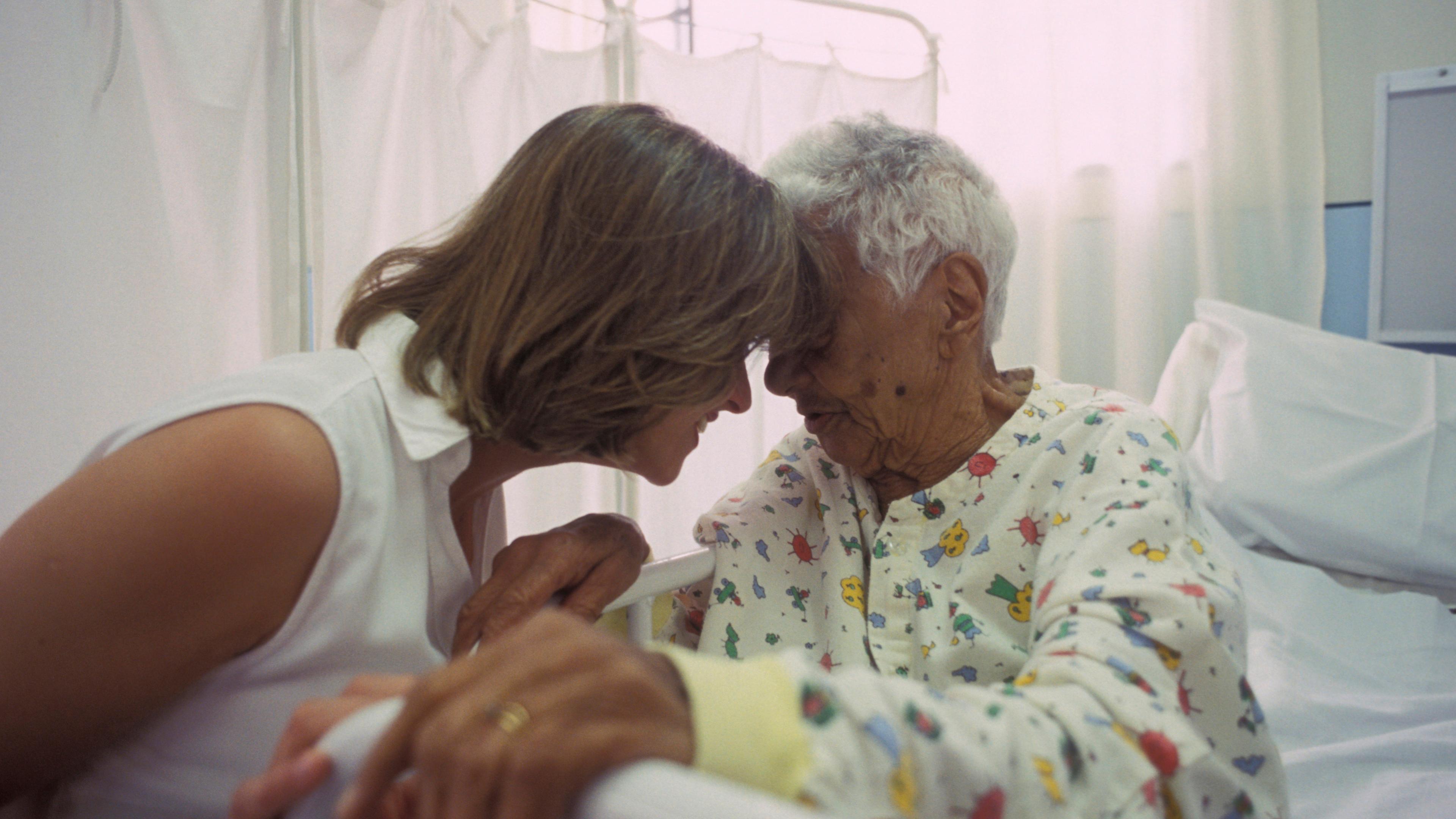 Photo of a woman in white comforting an elderly woman in a hospital gown with their foreheads touching in a hospital room.
