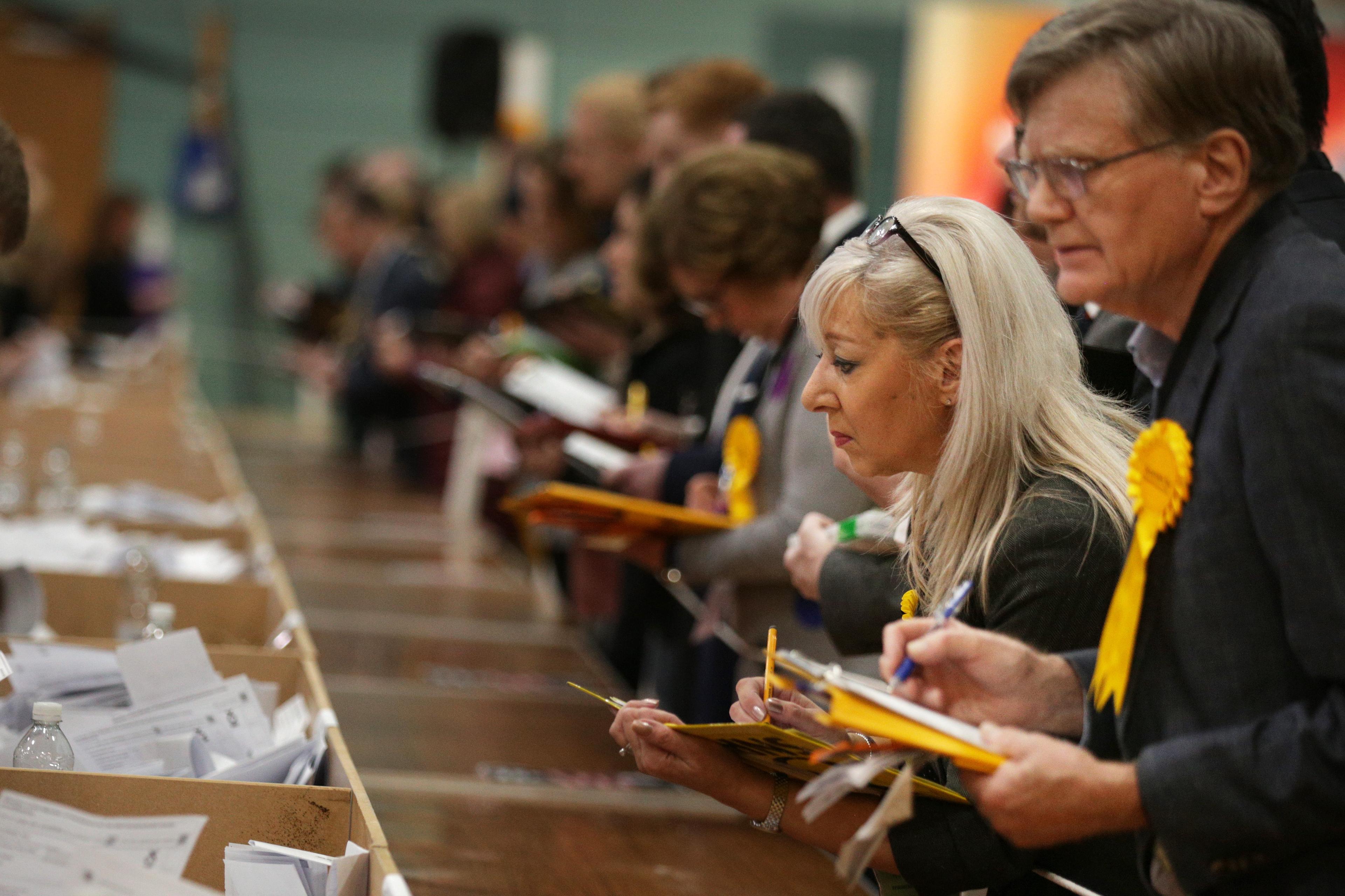 Photo of people counting ballots at an election table, wearing yellow rosettes, with focus on a woman and a man in the foreground.