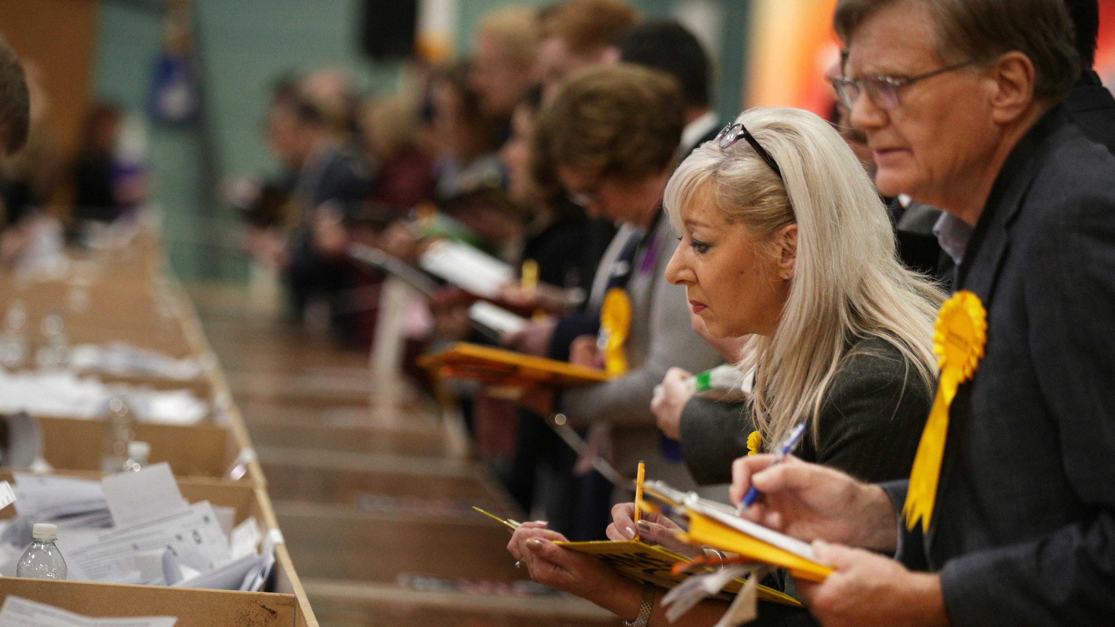 Photo of people counting ballots at an election table, wearing yellow rosettes, with focus on a woman and a man in the foreground.