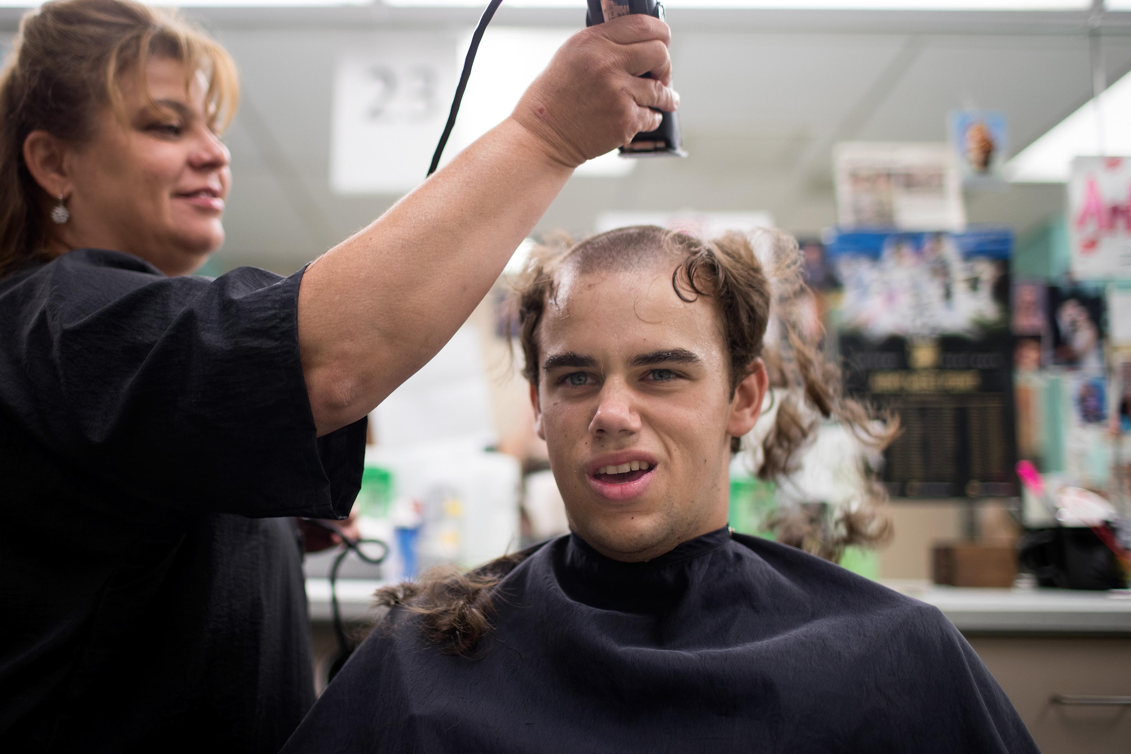 Photo of a boy getting a haircut by a hairdresser, hair partially shaved, grimacing expression, in a busy salon.