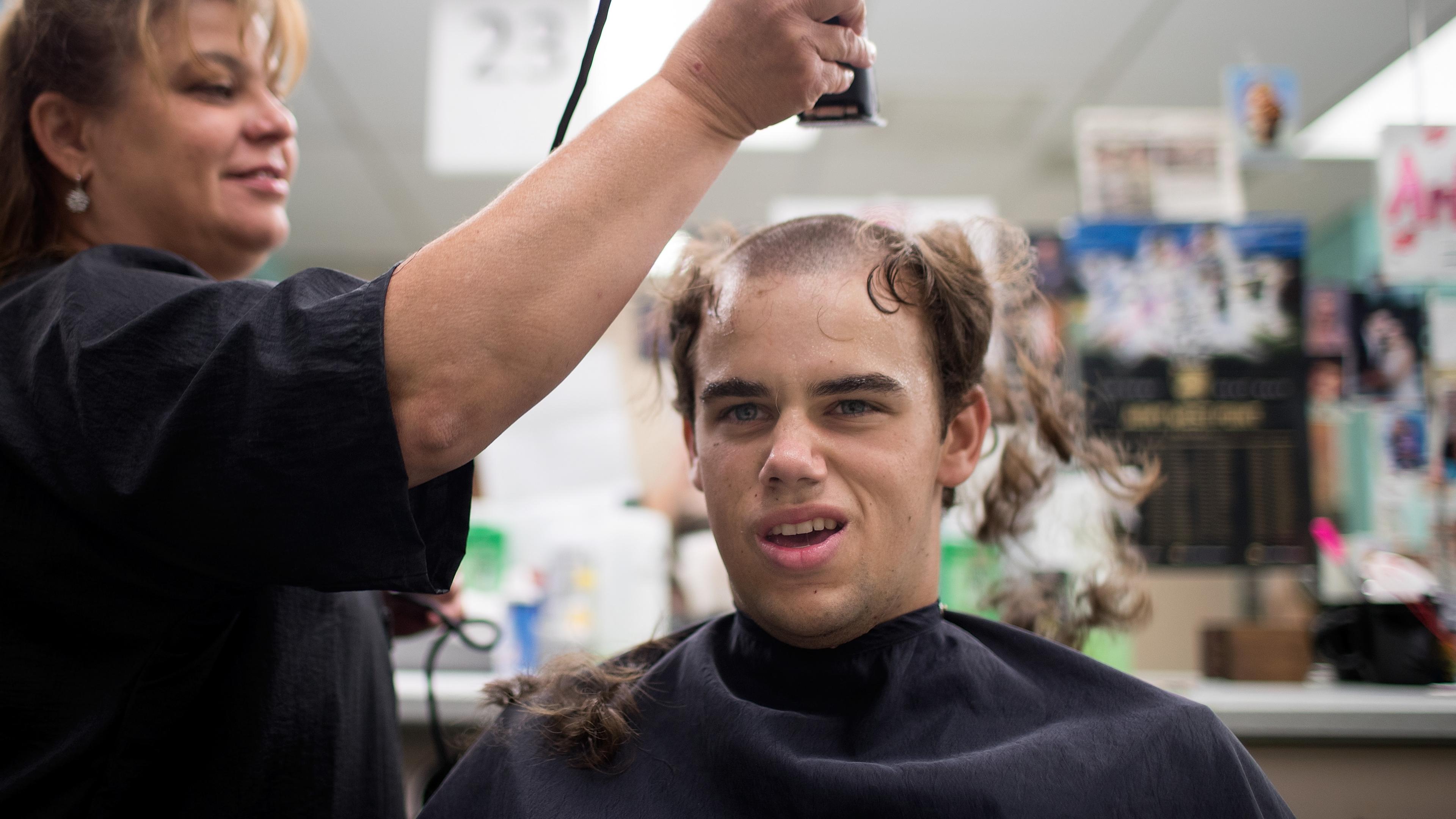 Photo of a boy getting a haircut by a hairdresser, hair partially shaved, grimacing expression, in a busy salon.